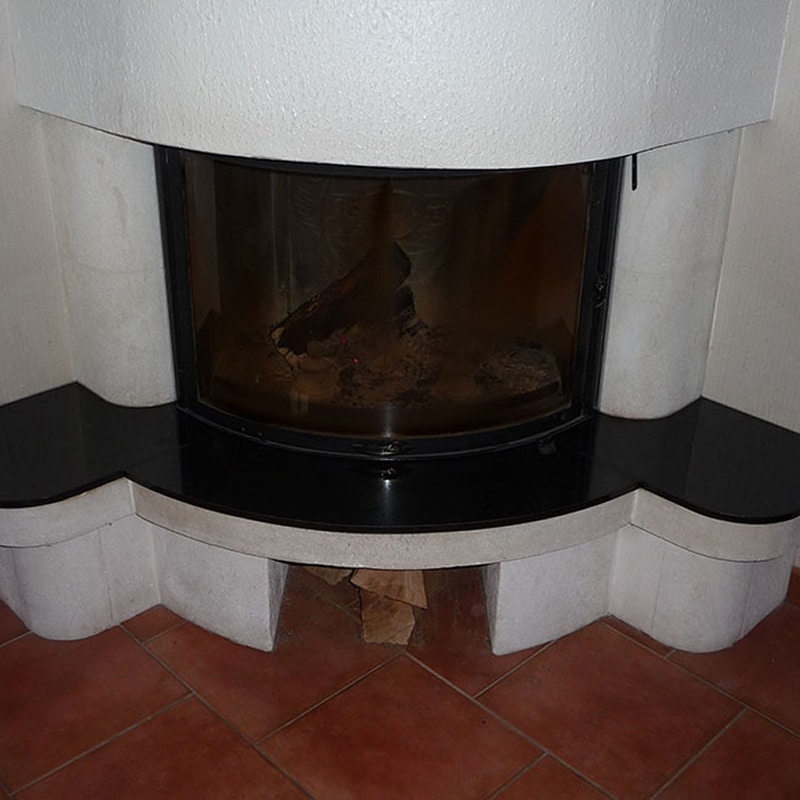 Cheminée moderne encastrée blanche avec insert vitré et braises. Tablette noire brillante, rangement à bûches intégré. Sol en terre cuite.