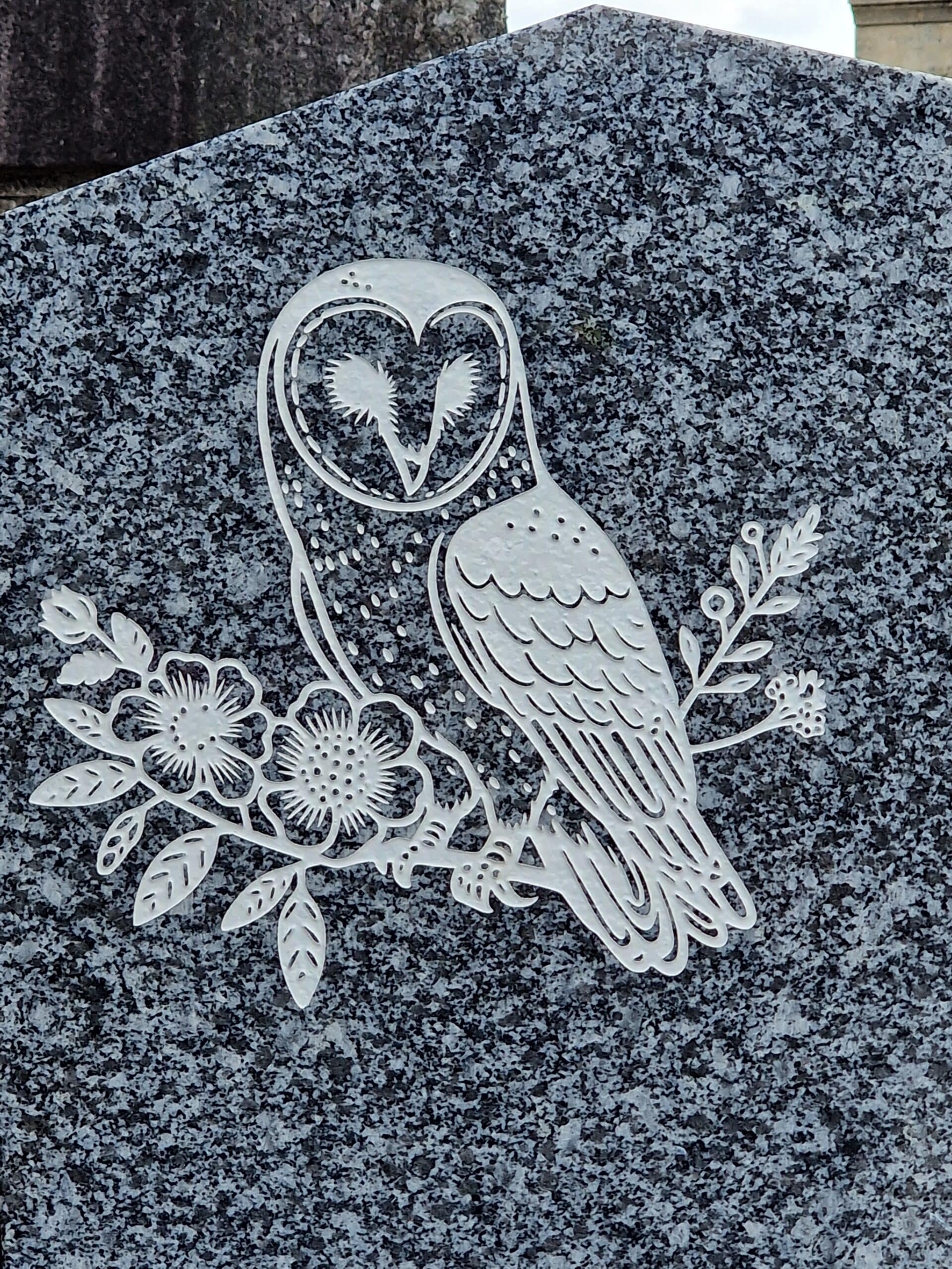 Chouette effraie gravée en blanc sur une branche de fleurs, ciselée dans une pierre tombale en granit gris moucheté.