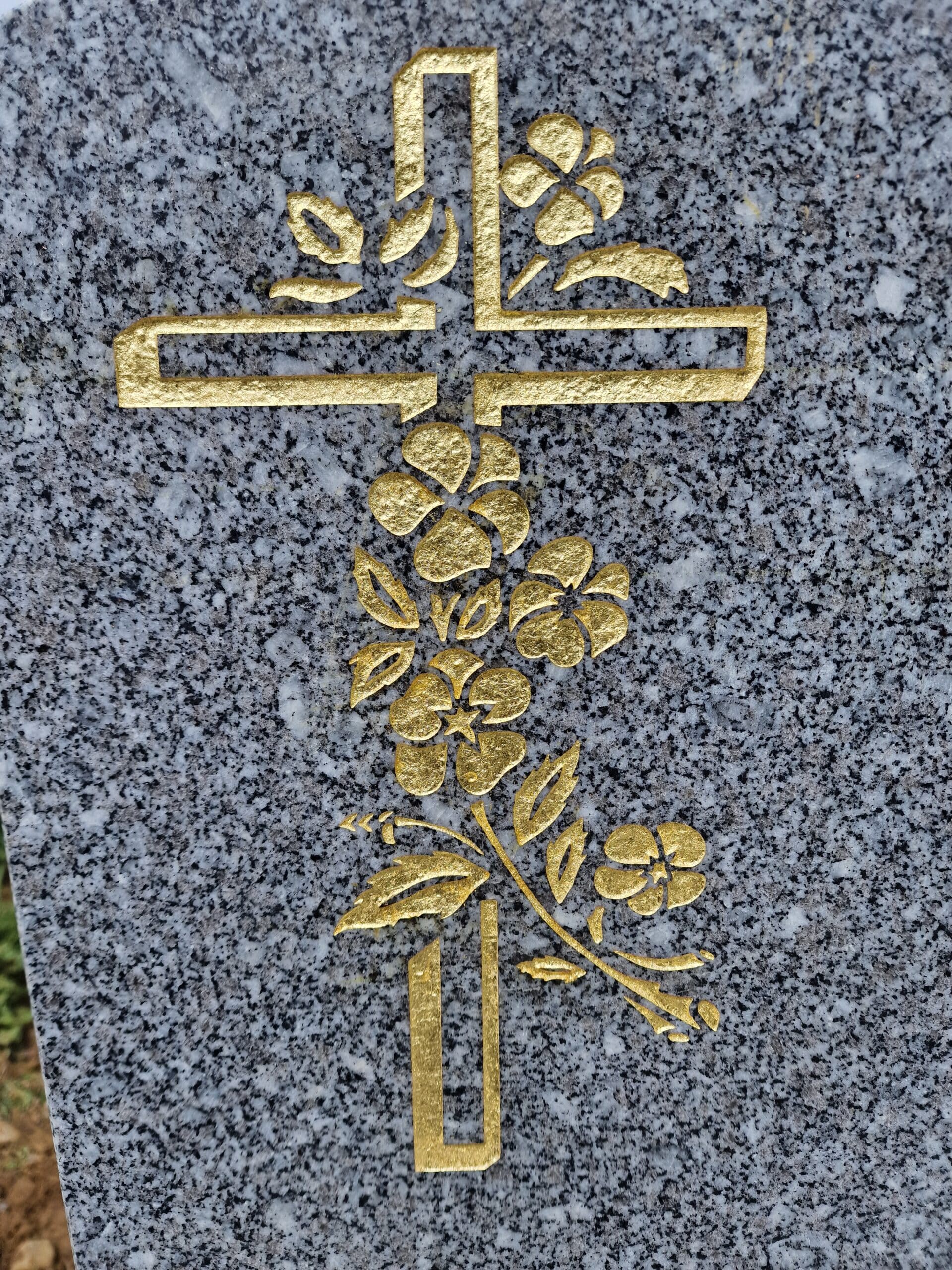 Croix dorée ornée de fleurs gravée sur une stèle en granit gris moucheté, symbole de paix et de recueillement.