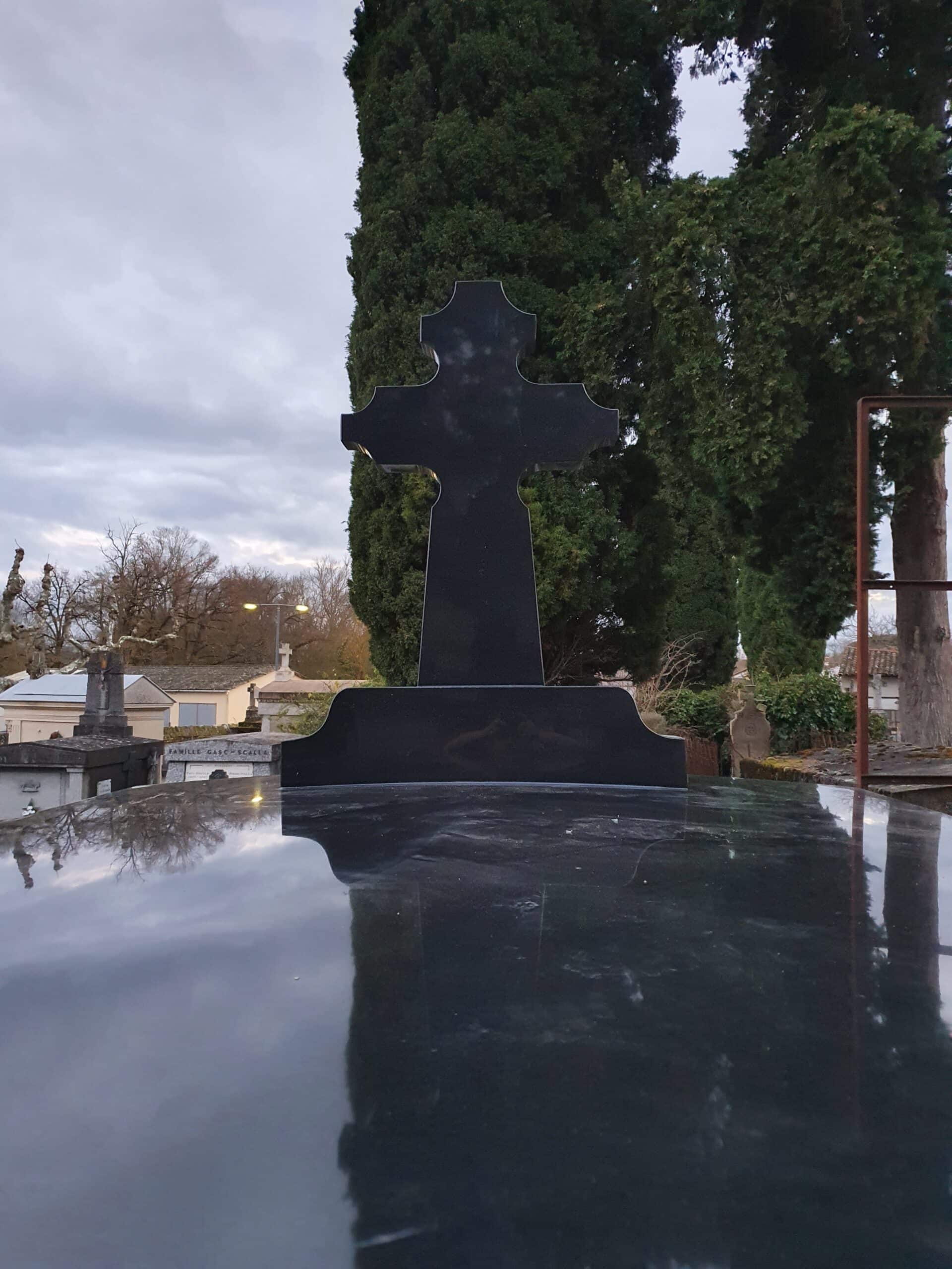Croix en granit noir poli avec reflet sur une stèle funéraire dans un cimetière arboré.