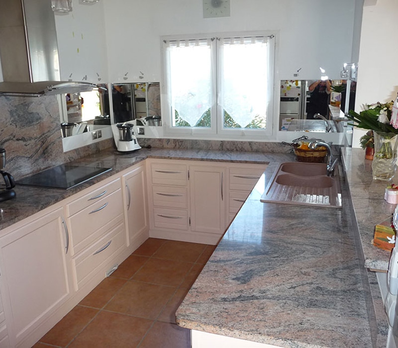 Cuisine moderne en U avec comptoirs en granit gris-rose, meubles beiges et carrelage terre cuite. Évier double, hotte, fenêtres lumineuses.