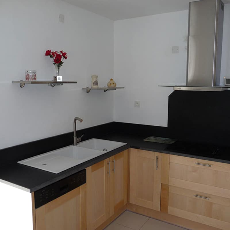 Cuisine moderne avec meubles en bois clair, comptoir noir et évier double. Étagères murales décorées et hotte inox au-dessus d'une plaque de cuisson.