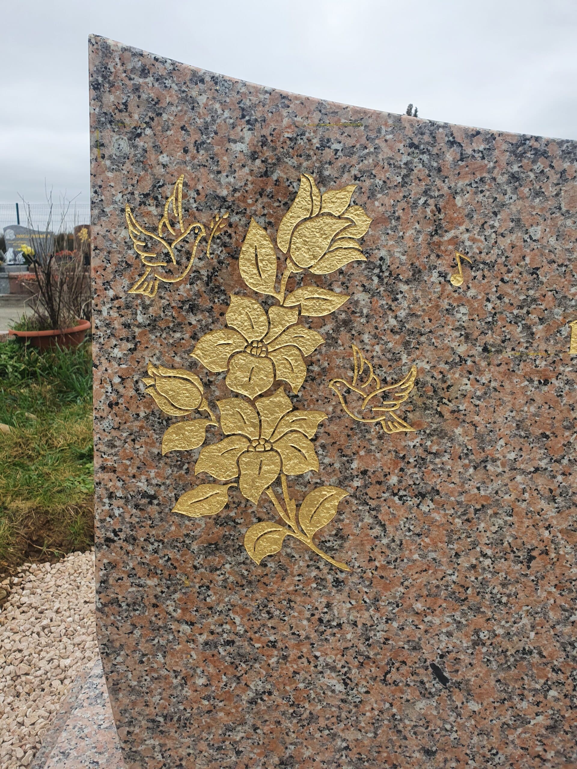 Motifs dorés de fleurs, oiseaux et note de musique gravés sur une pierre tombale en granit rose moucheté.