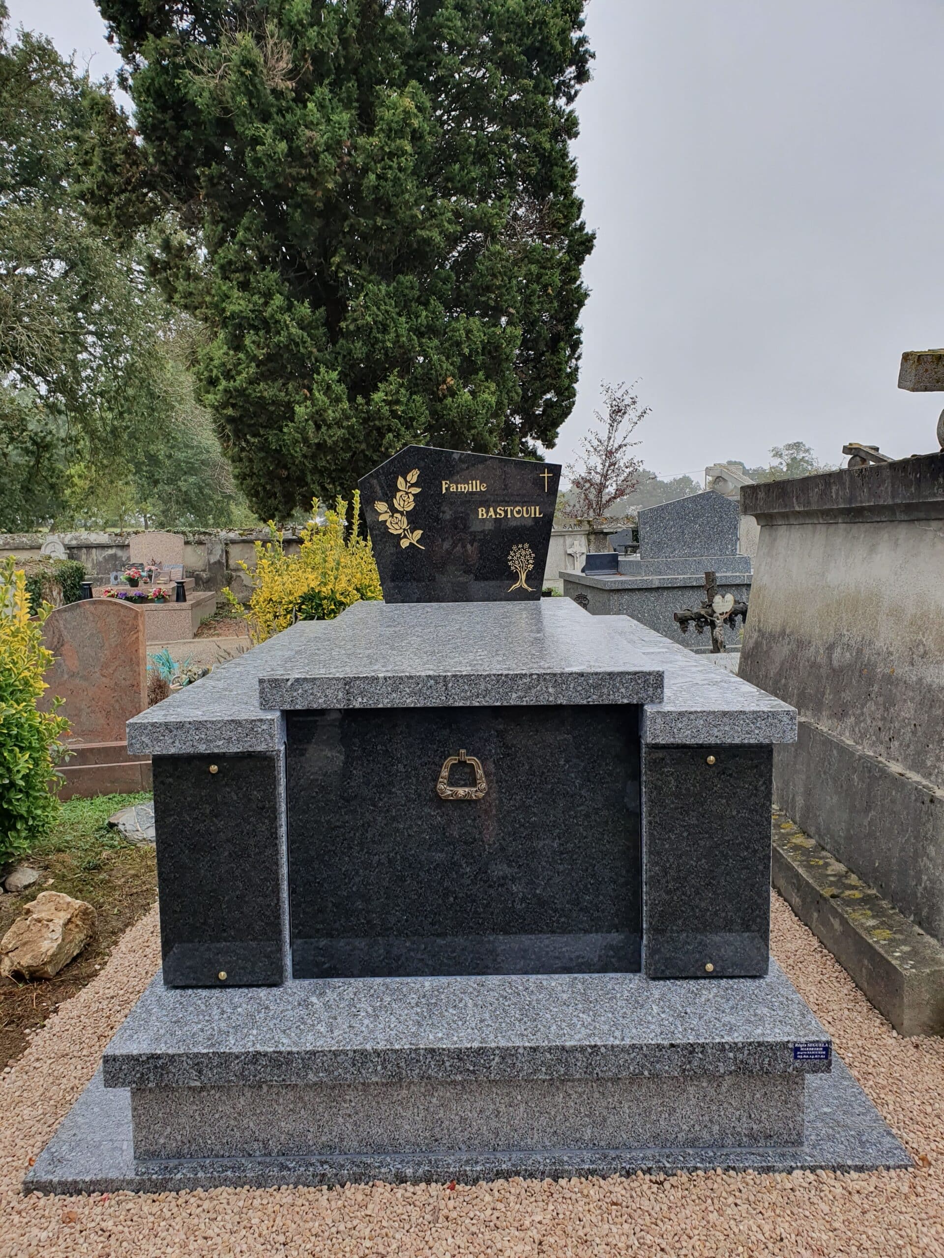 Monument funéraire en granit gris et noir de la Famille BASTOUIL avec gravures dorées dans un cimetière verdoyant.