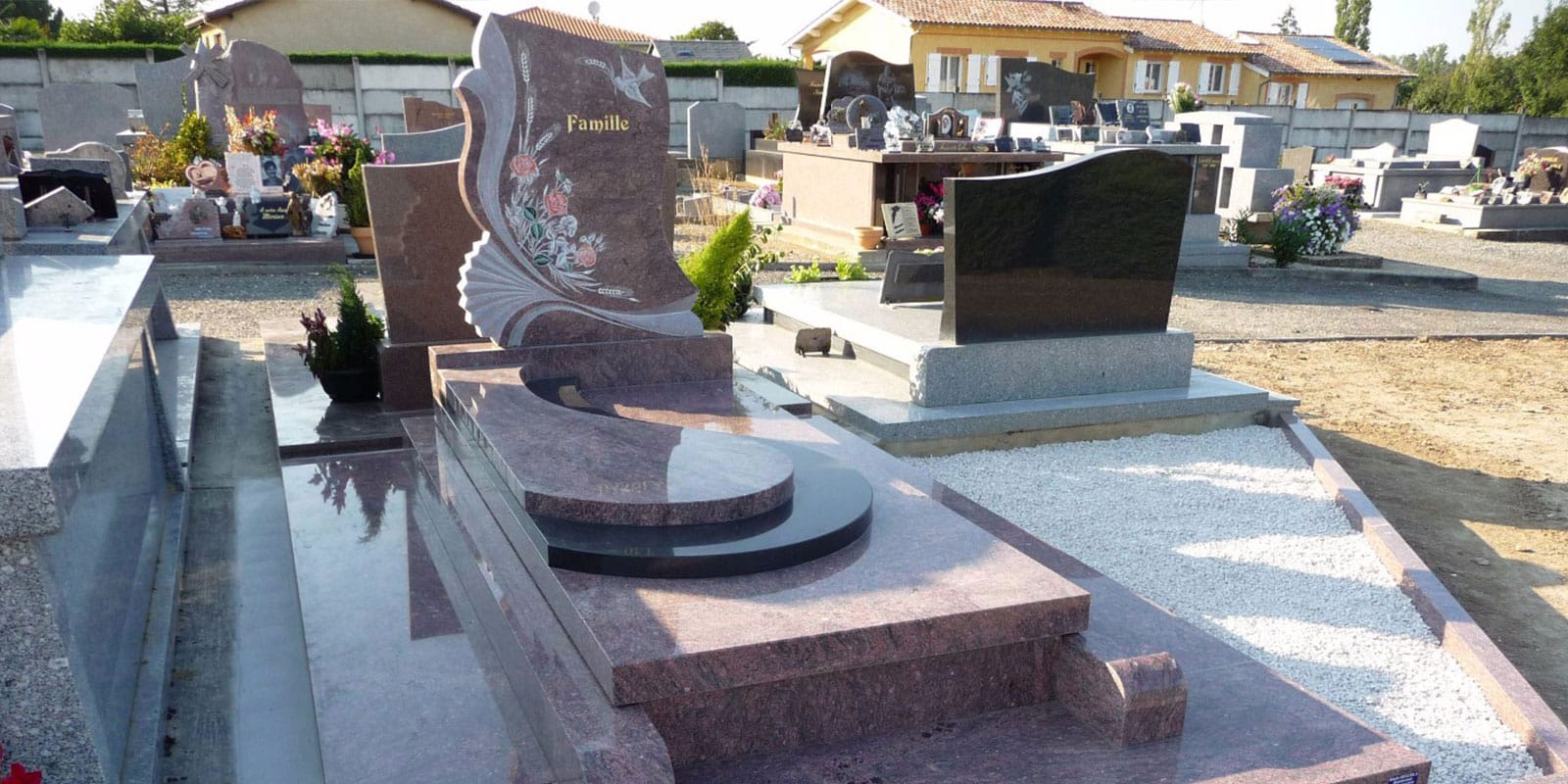 Cimetière ensoleillé avec un monument funéraire en granite rose et noir, sculpté "Famille" avec des motifs floraux et un oiseau. D'autres tombes et une maison jaune sont visibles en arrière-plan.