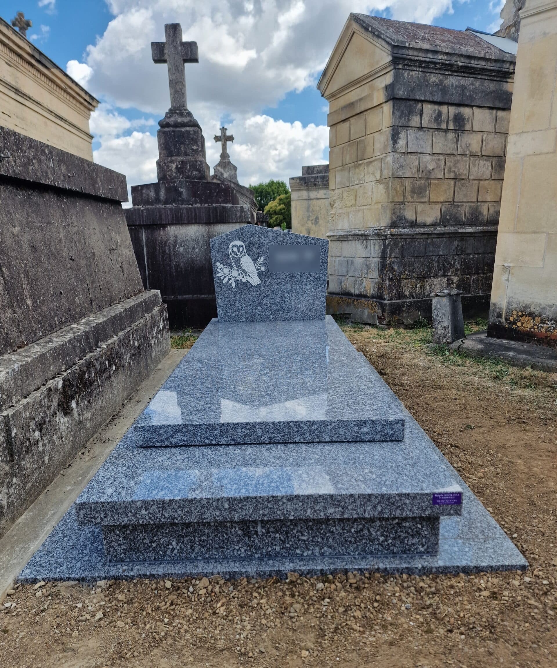 Monument funéraire en granit bleu poli avec gravure de chouette dans un cimetière sous un ciel nuageux.