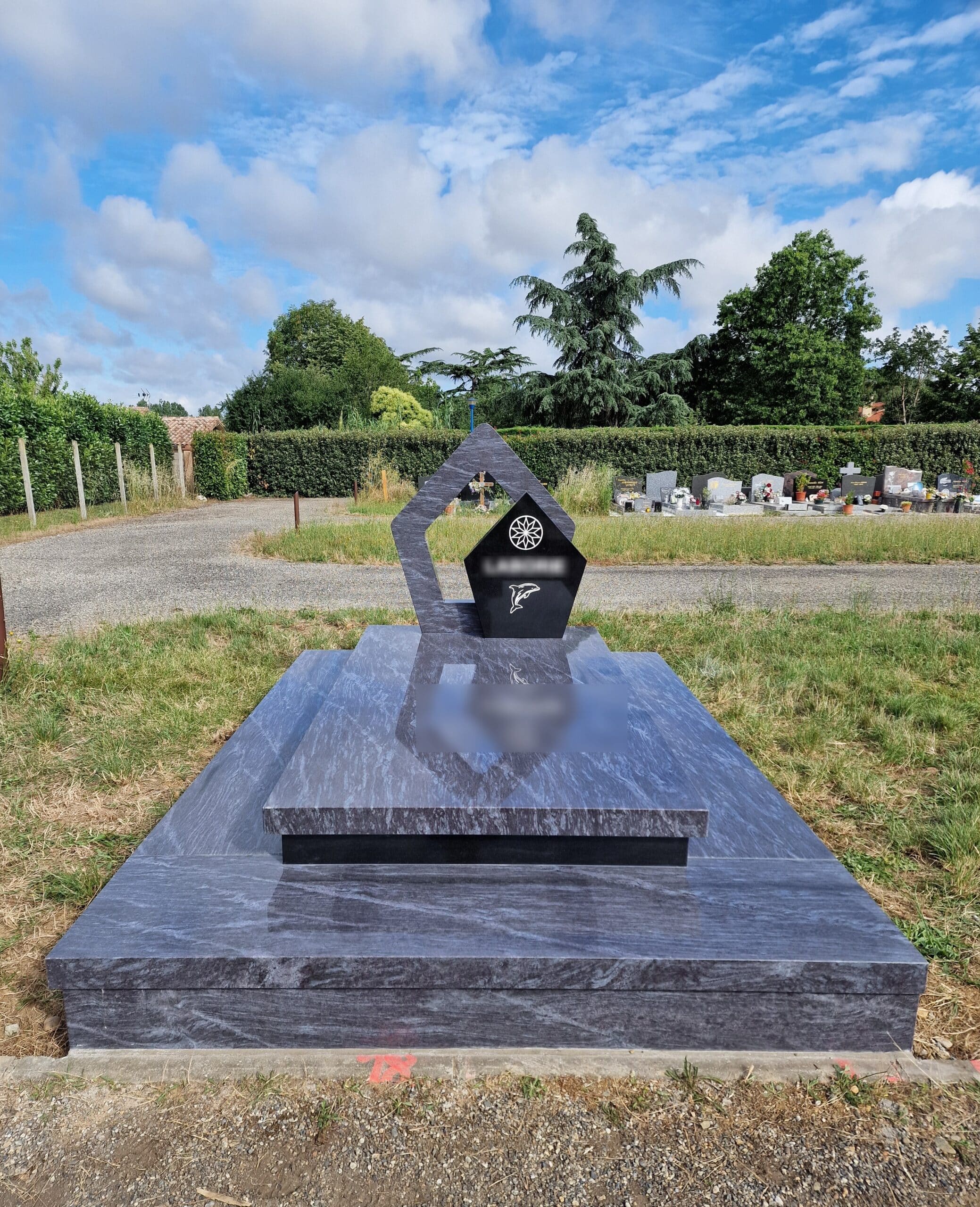 Monument funéraire moderne en granit gris bleuté, stèle géométrique avec dauphin, dans un cimetière paysagé.