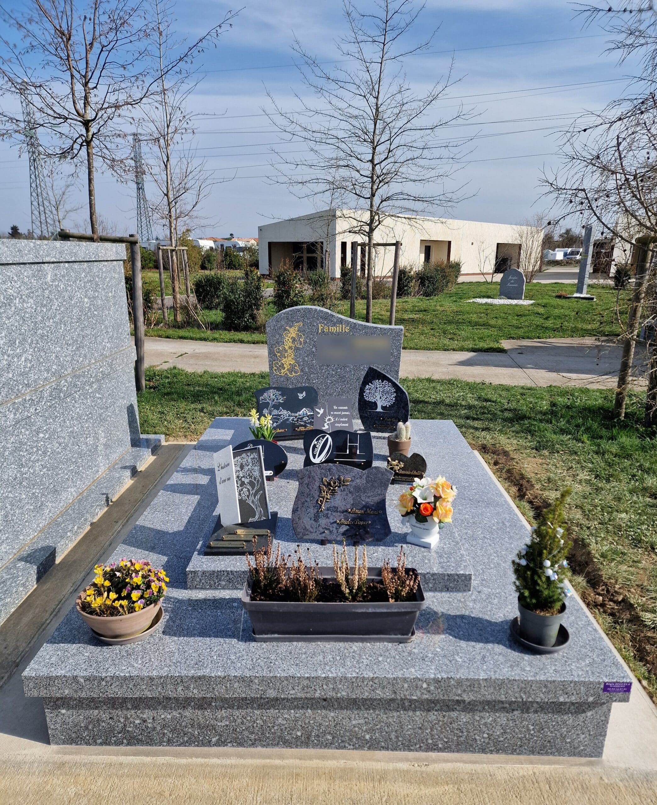 Monument funéraire en granit gris avec plaques, fleurs et gravure 'Famille' au cœur d'un cimetière paysager moderne.
