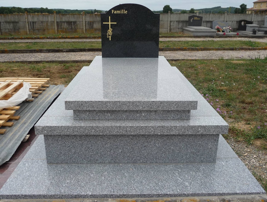 Monument funéraire moderne en granit poli gris clair, avec stèle noire gravée 'Famille' et croix dorée. Environnement cimetière.