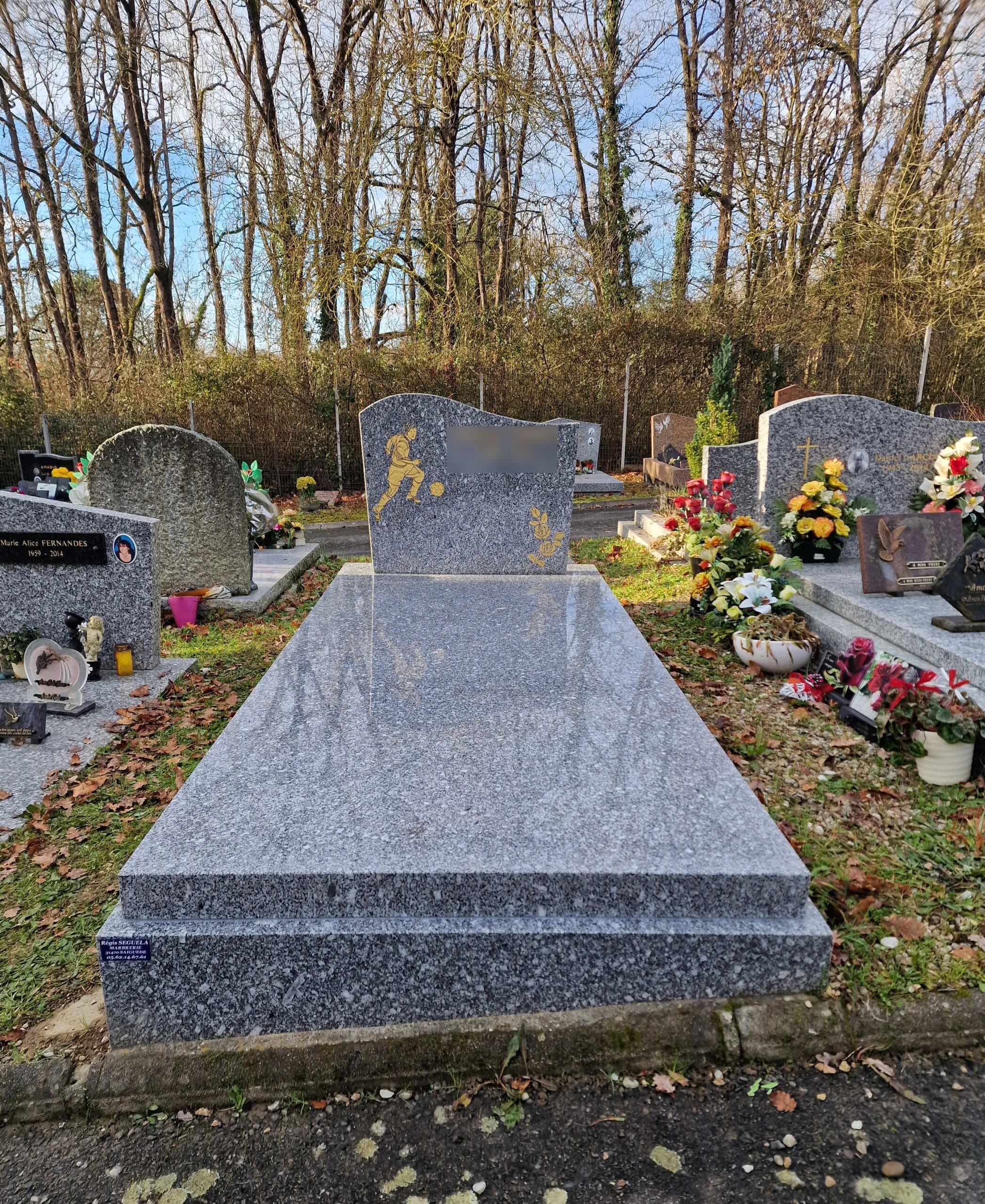 Monument funéraire en granit gris avec gravure dorée d'un footballeur et d'une rose dans un cimetière boisé.