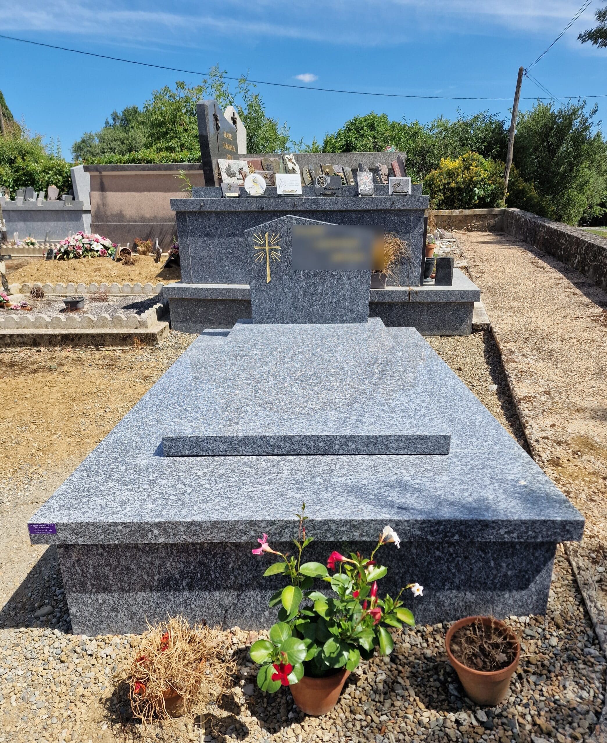 Monument funéraire en granit gris poli avec des fleurs dans un cimetière sous un grand ciel bleu.
