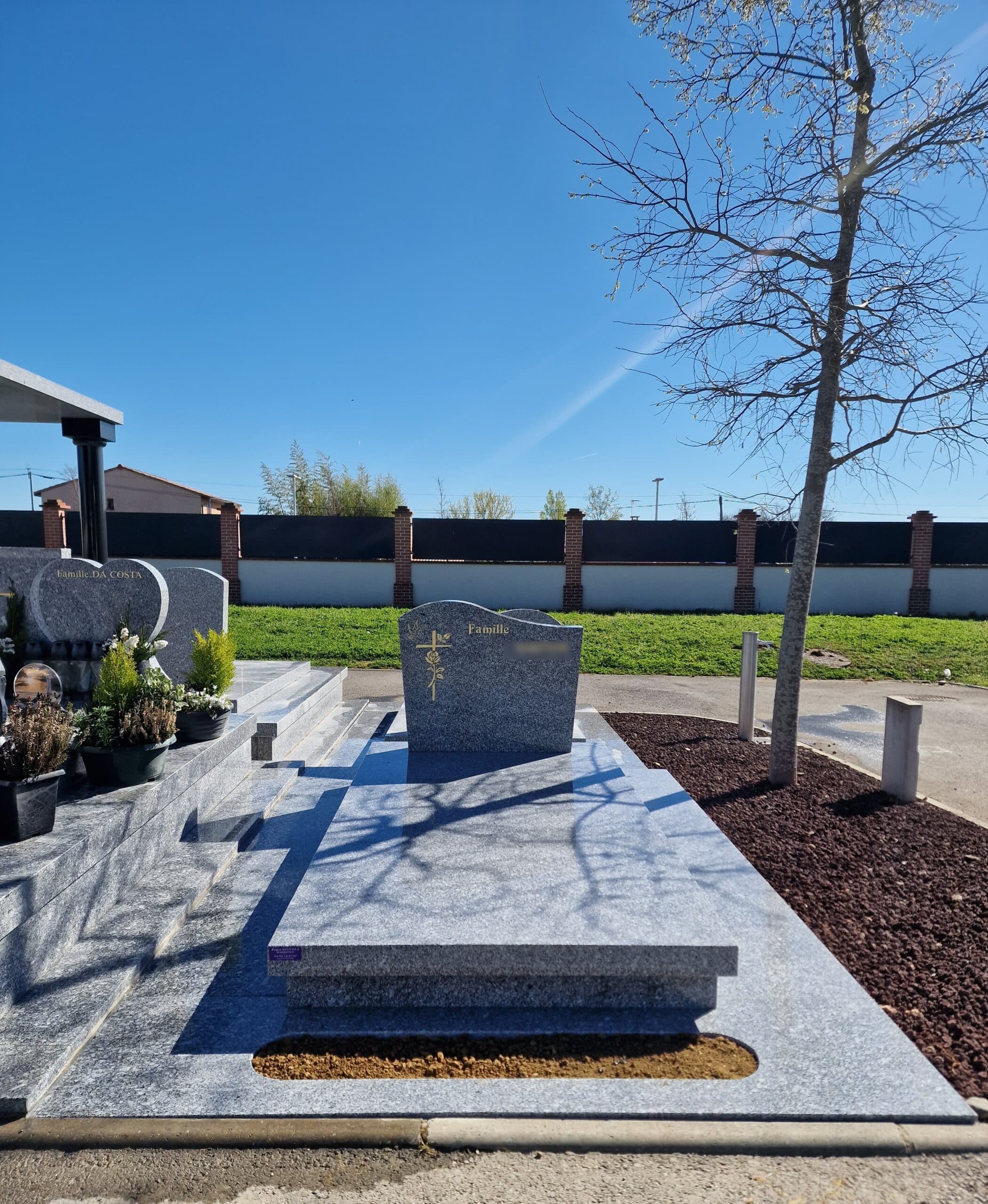 Monument funéraire en granit gris avec croix dorée et inscription Famille sous un ciel bleu dégagé.