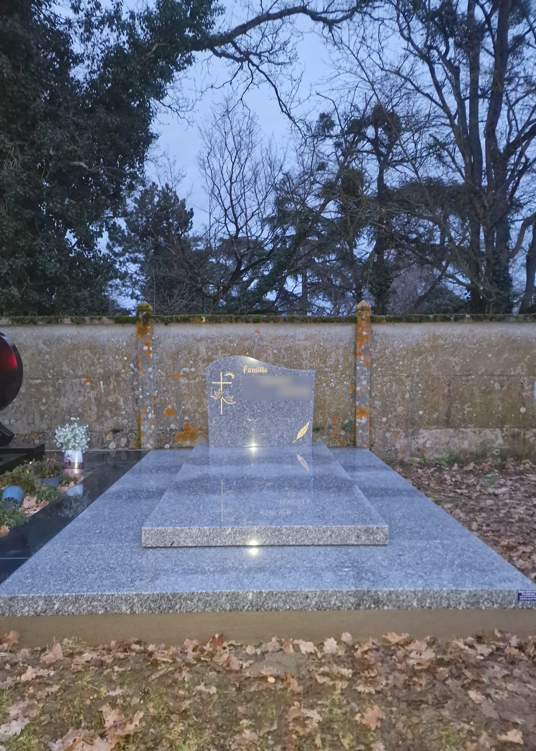 Tombeau en granit gris avec stèle gravée Famille et croix dorée devant un mur de pierre et des arbres dépouillés.