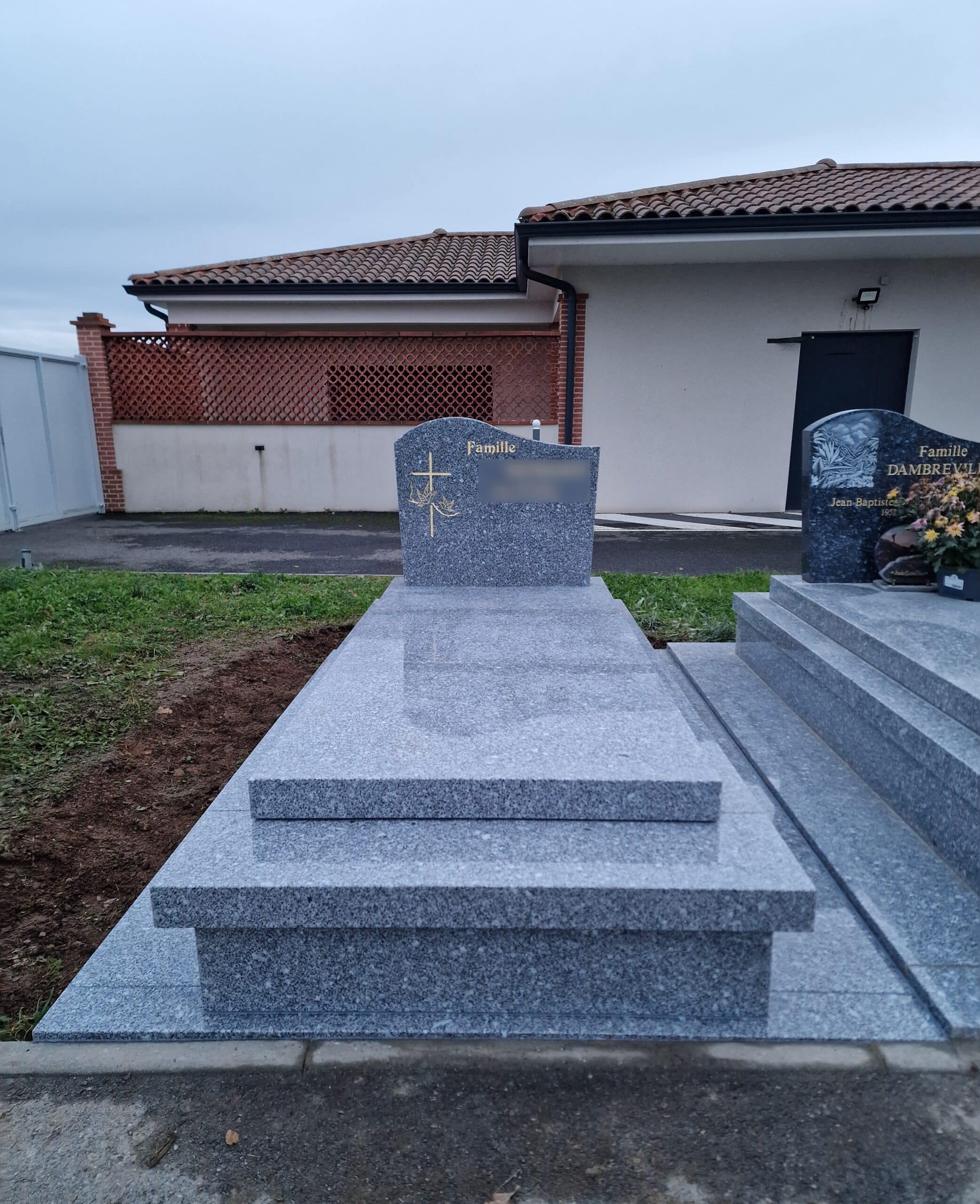 Monument funéraire en granit gris avec croix dorée et mention Famille devant un bâtiment moderne.