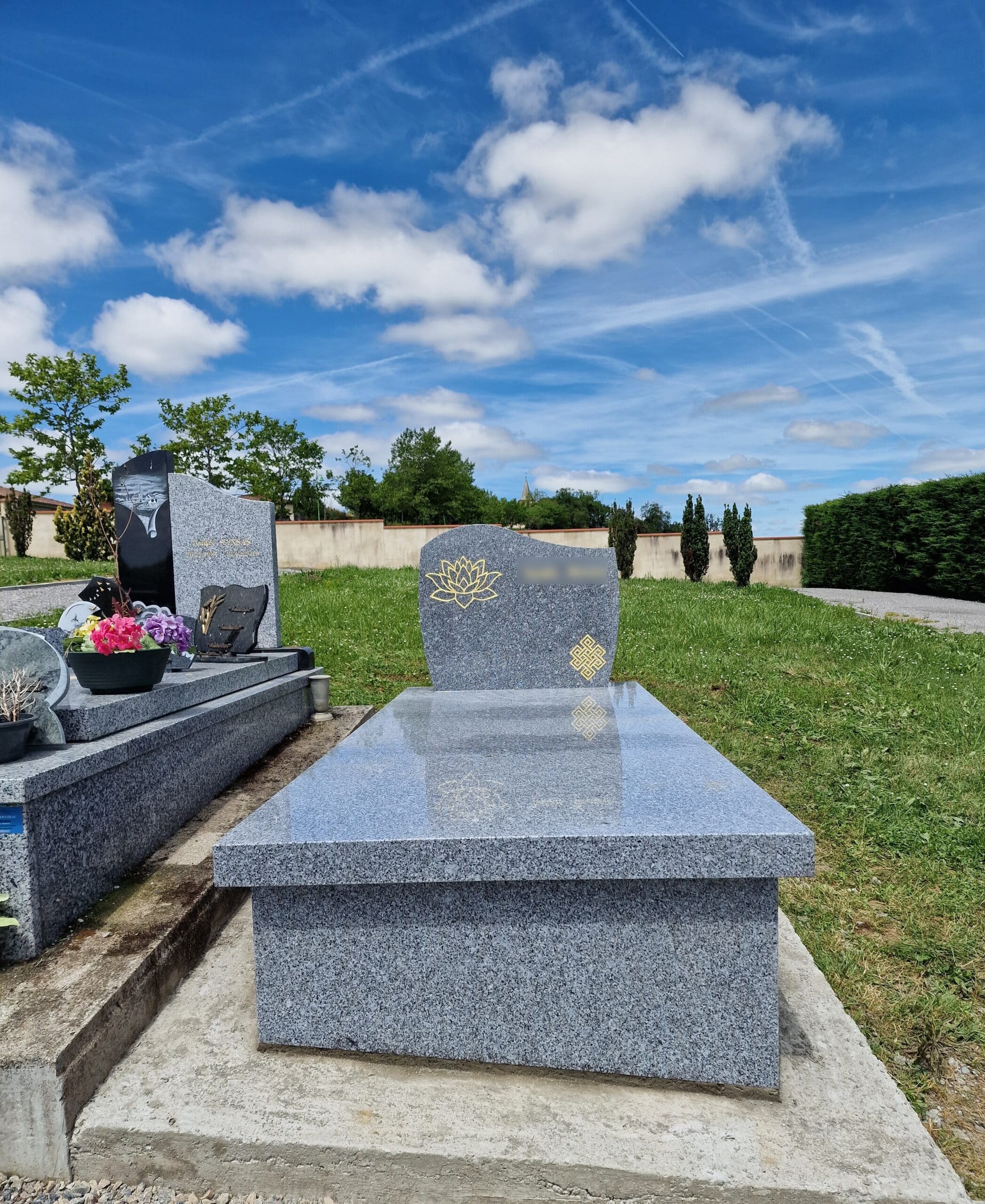 Pierre tombale en granit gris avec lotus doré et nœud éternel sous un ciel bleu nuageux dans un cimetière paysager.