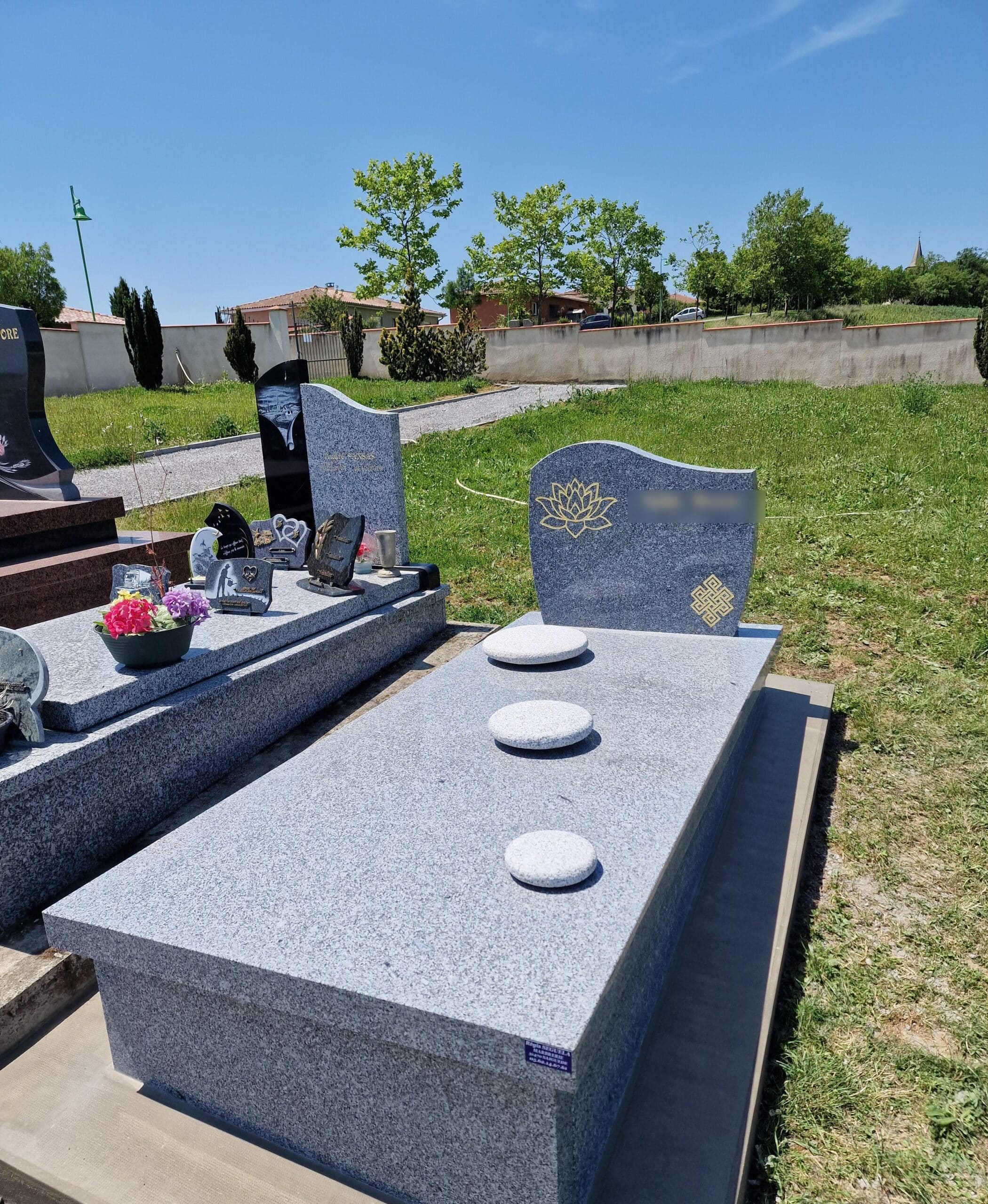 Monuments funéraires modernes en granit gris avec symboles de lotus et nœud éternel sous un ciel bleu dégagé.