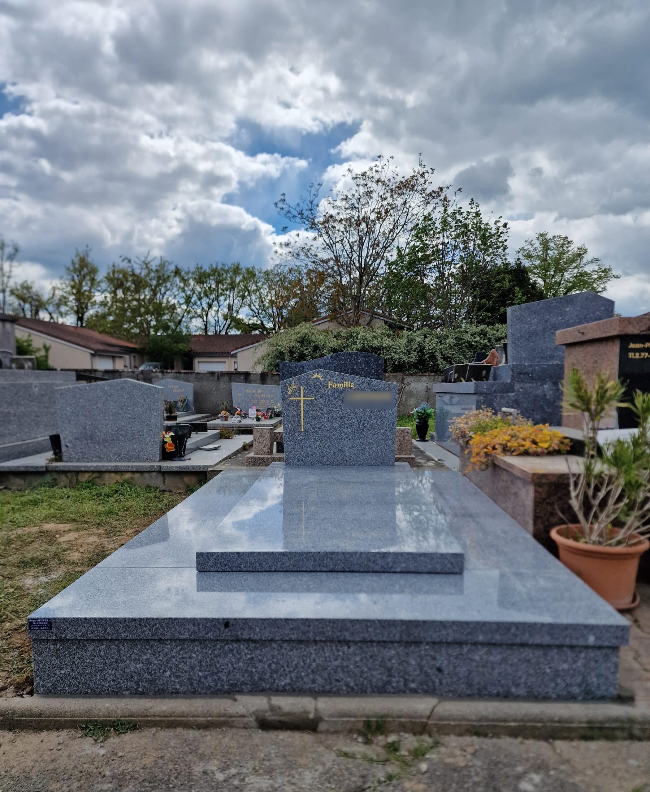 Monument funéraire en granit gris poli avec une croix dorée sous un ciel nuageux dans un cimetière paysager.