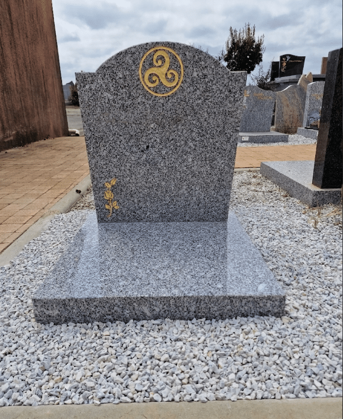Monument funéraire en granit gris avec triskèle doré et rose, entouré de graviers blancs.