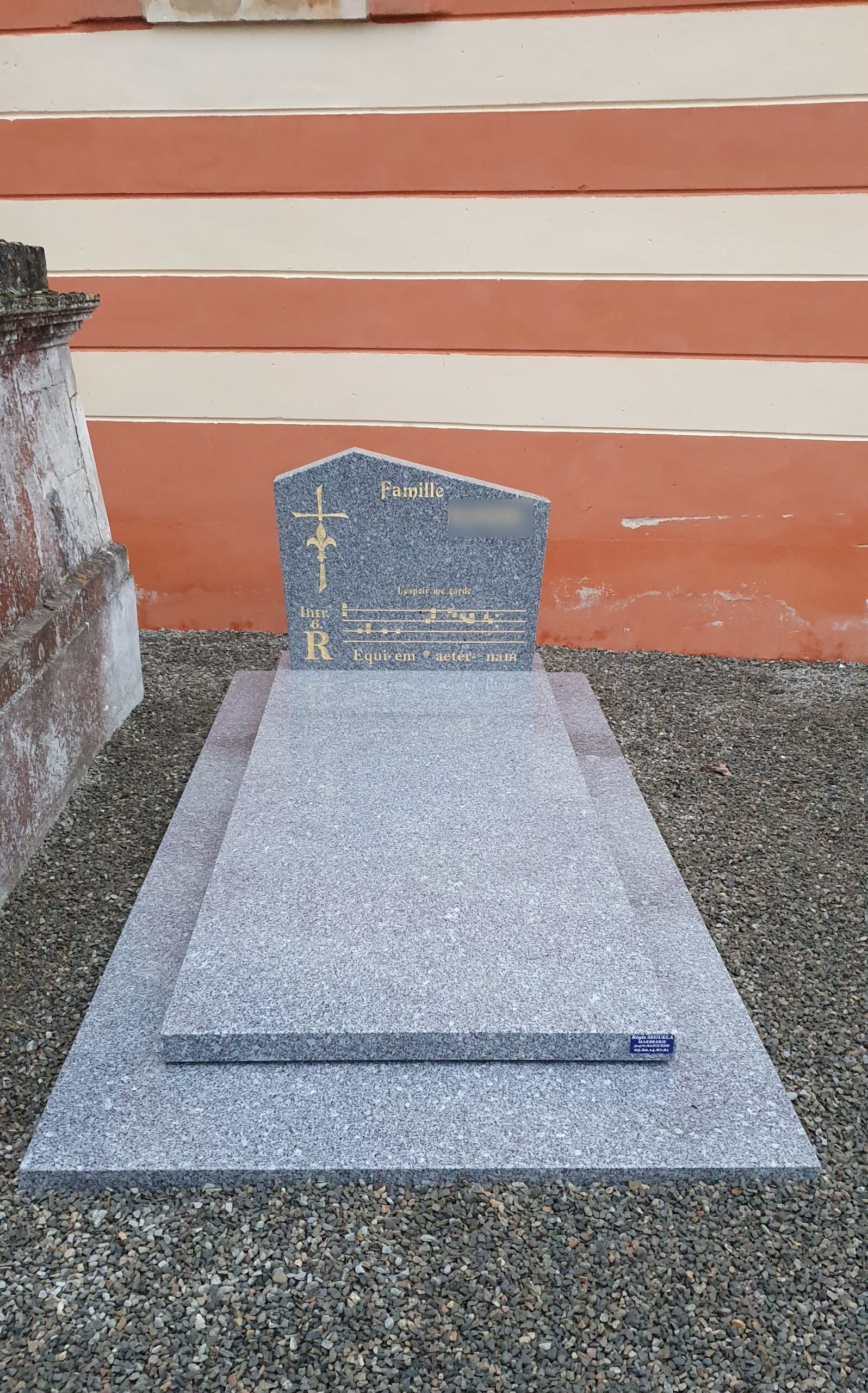 Tombeau en granit gris avec partition de Requiem gravée, croix fleurdelisée et inscription Famille, devant mur orange.