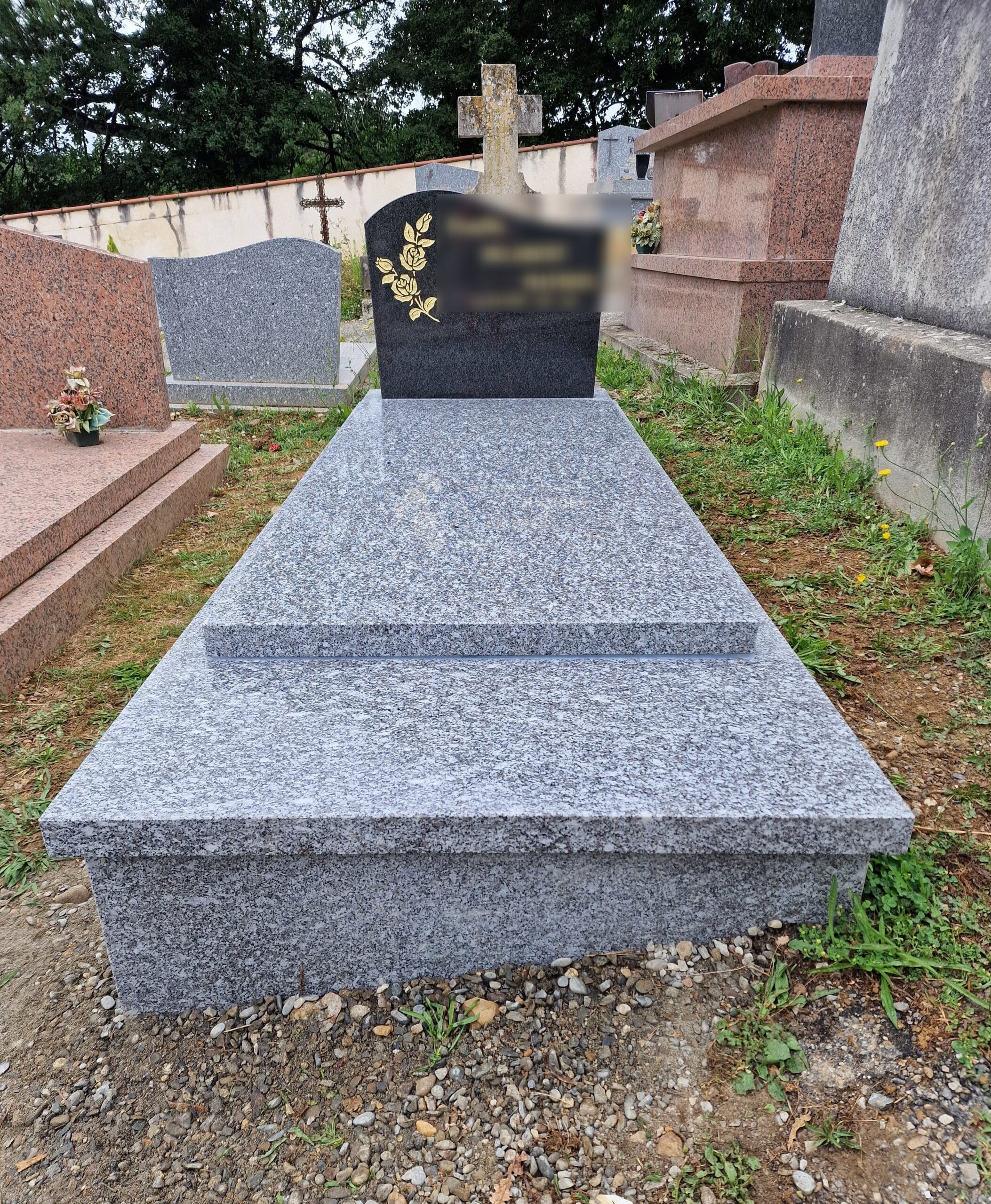 Monument funéraire en granit gris avec stèle noire ornée d'une rose dorée et croix en pierre dans un cimetière.