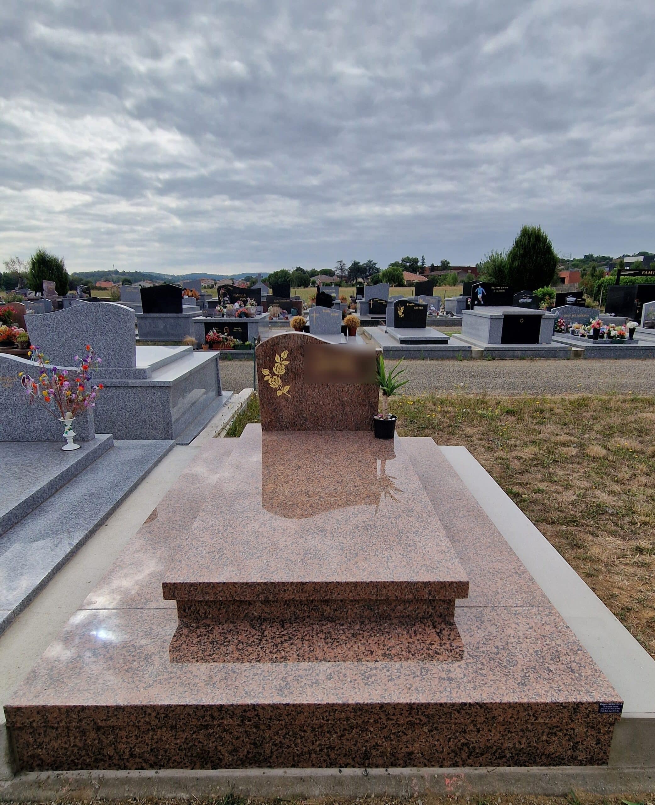 Monument funéraire en granit rose avec une rose dorée gravée dans un cimetière sous un ciel nuageux.
