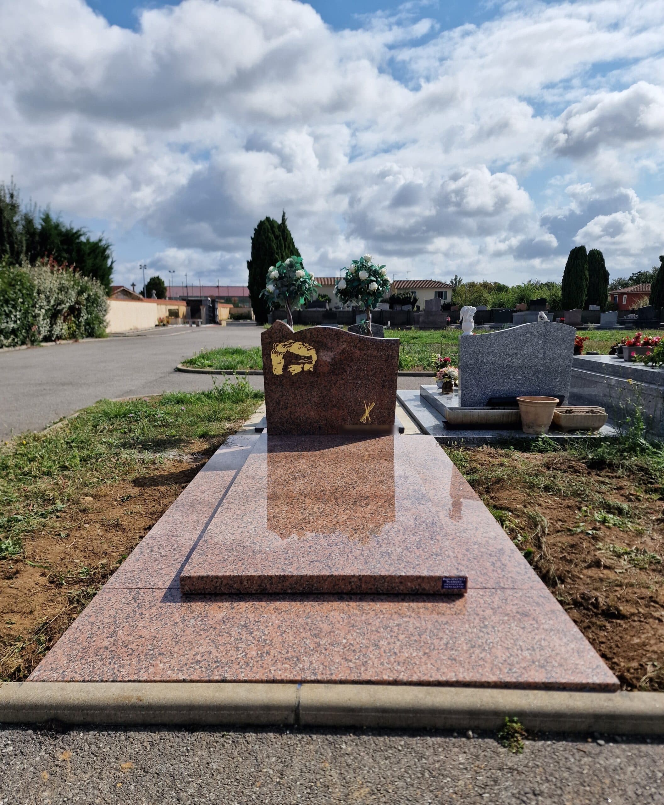 Monument funéraire en granit rose avec gravure dorée de ciseaux dans un cimetière paysager sous un ciel nuageux.