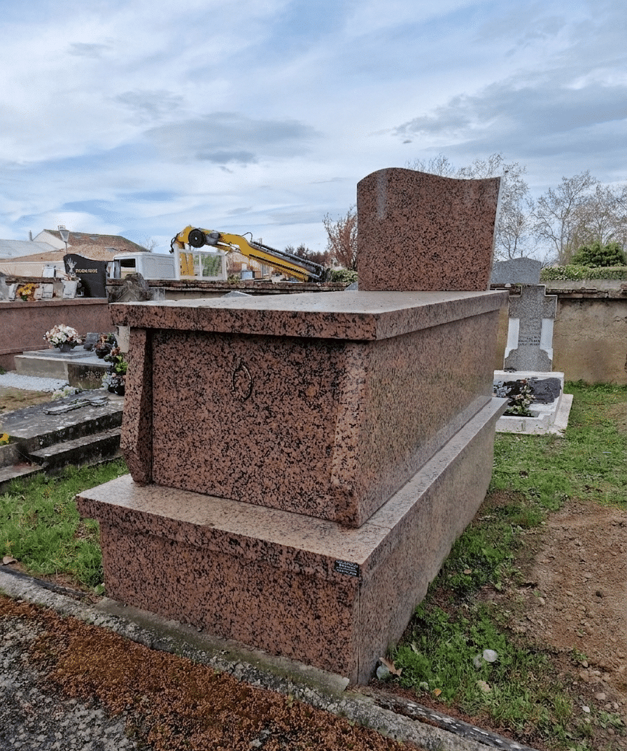Monument funéraire massif en granit rose moucheté dans un cimetière sous un ciel nuageux avec grue en arrière-plan.