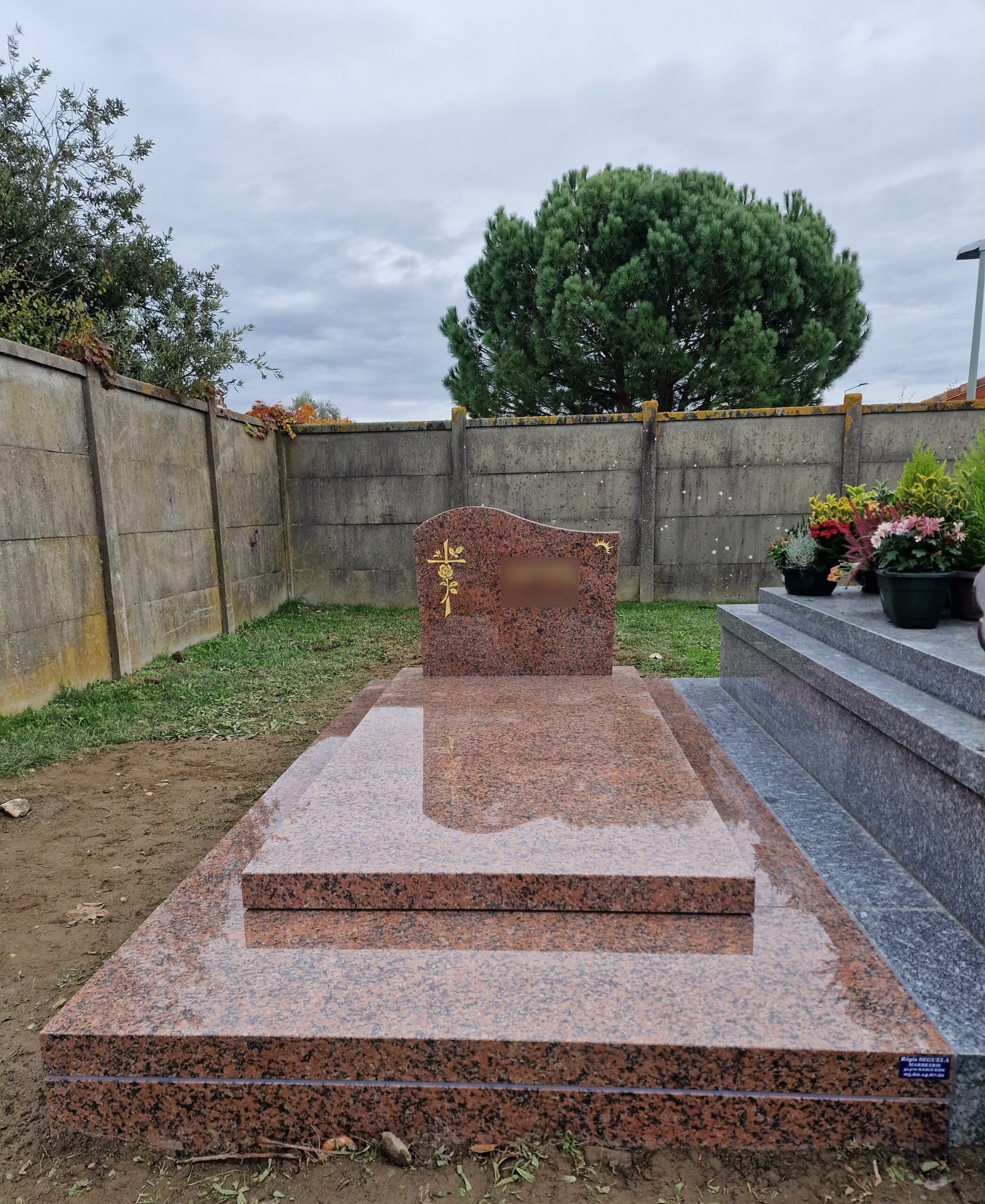 Monument funéraire en granit rouge poli avec croix et rose dorées, installé dans un cimetière calme sous un ciel nuageux.