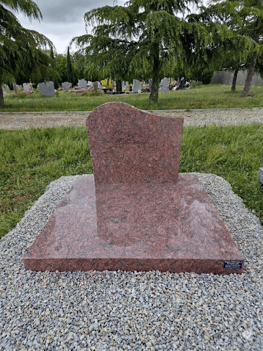 Monument funéraire en granit rouge et noir avec stèle arrondie sur socle en pierre dans un cimetière paysager.