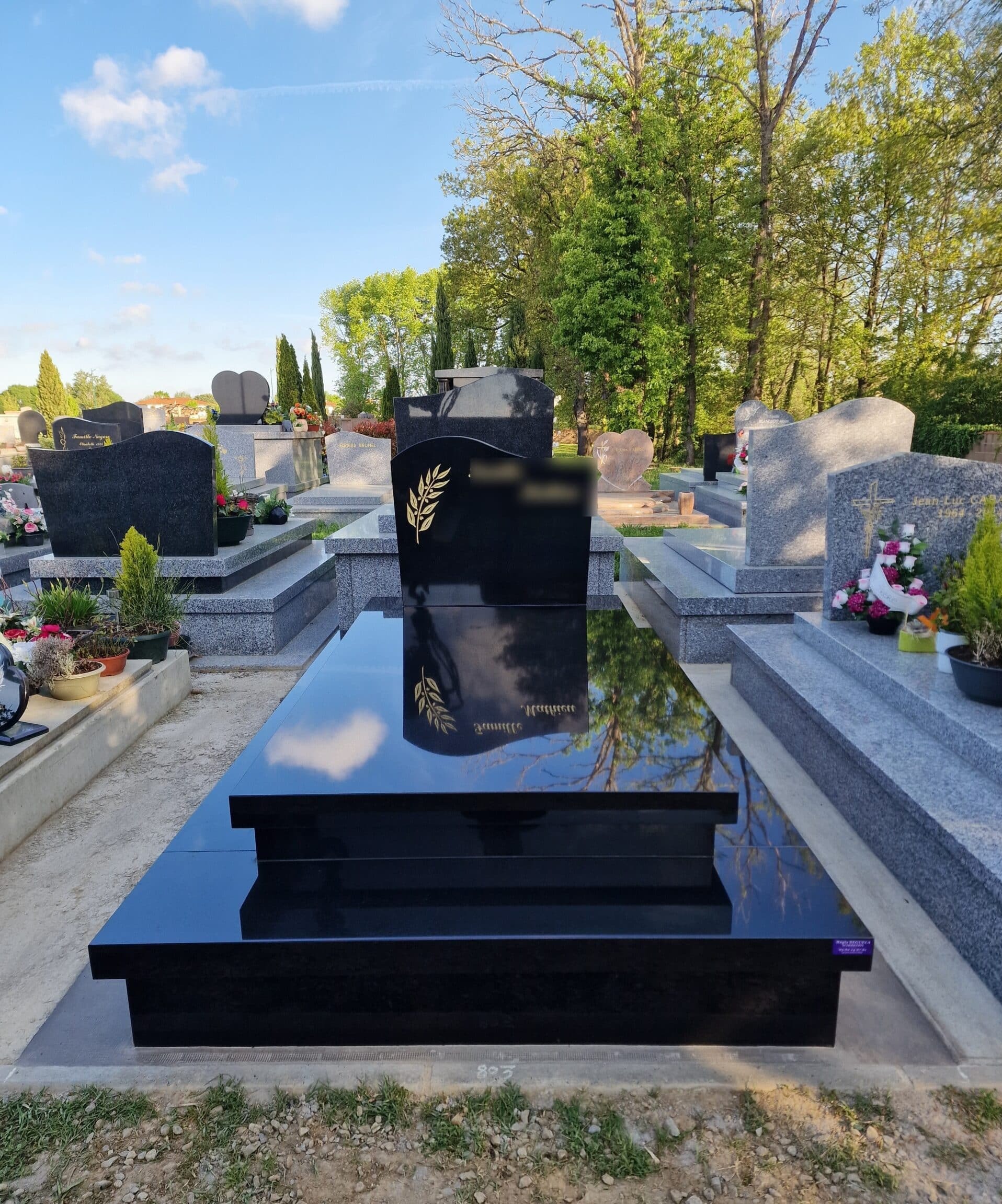 Pierre tombale moderne en granit noir poli avec épi de blé doré dans un cimetière paisible et arboré.