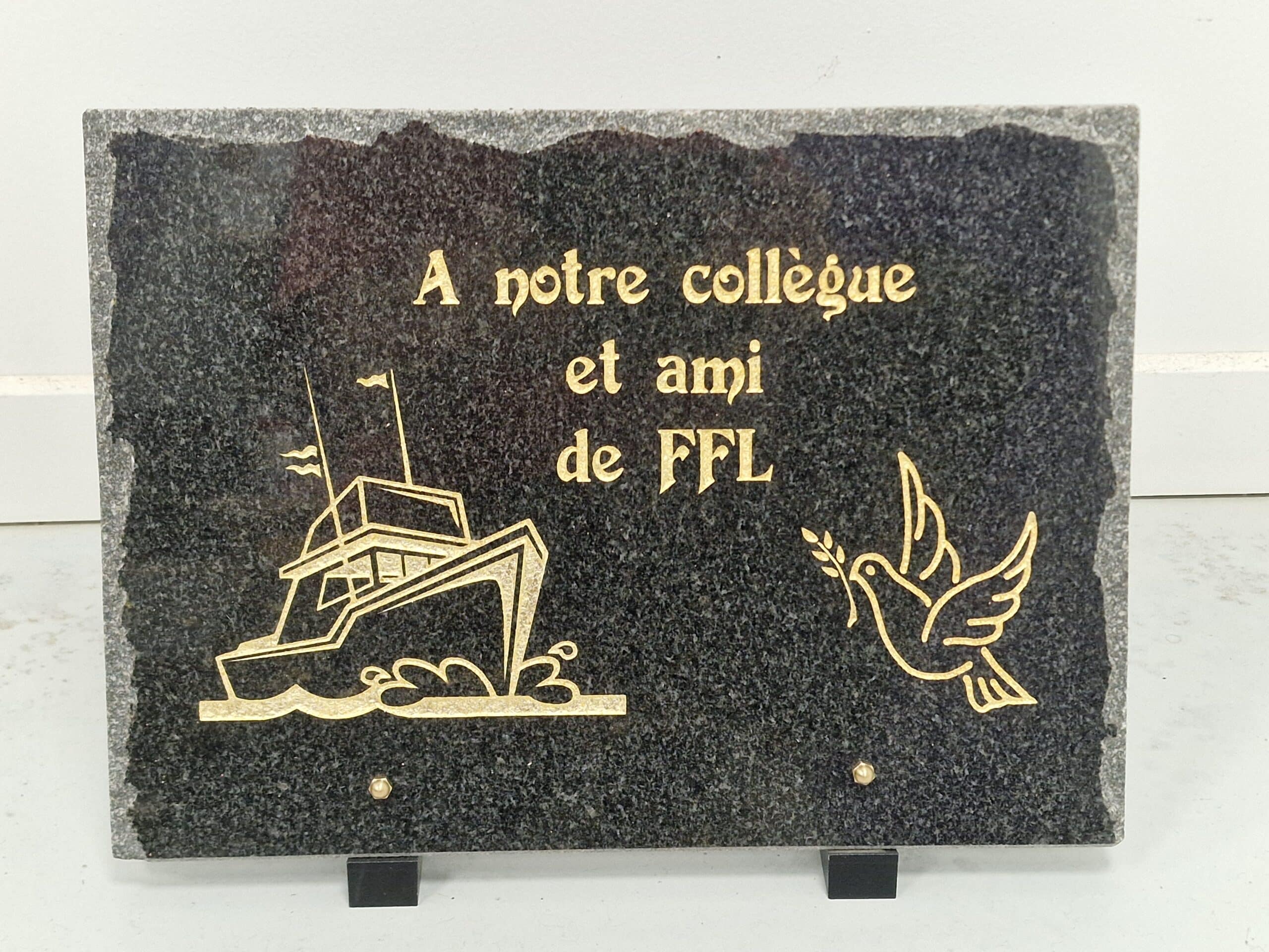 Plaque commémorative en granit noir gravée "A notre collègue et ami de FFL" avec un bateau et une colombe dorés.