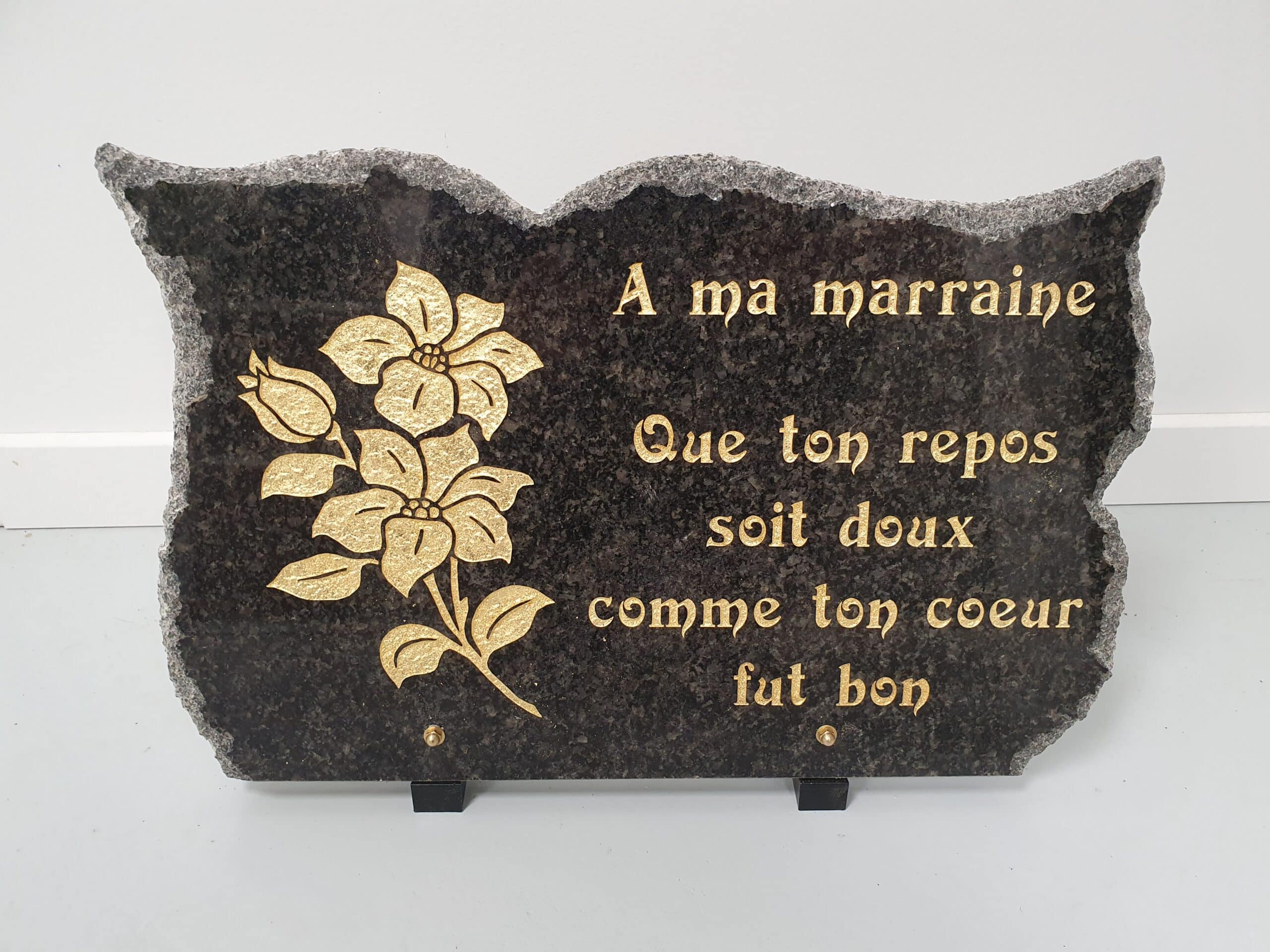 Plaque funéraire en granit noir avec fleurs dorées et texte : "A ma marraine Que ton repos soit doux comme ton coeur fut bon"