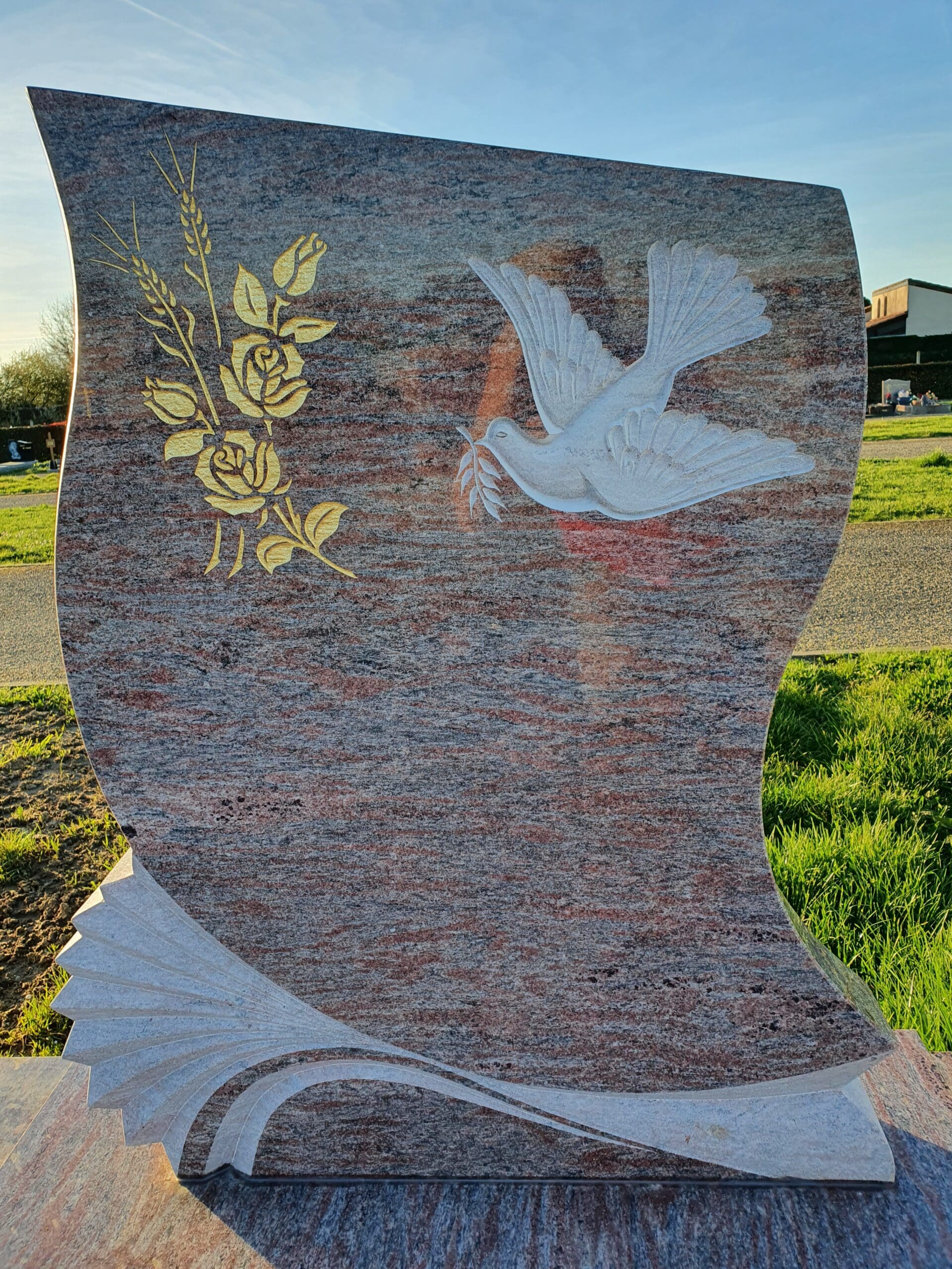 Stèle funéraire en granit rose avec colombe blanche sculptée et bouquet de roses dorées dans un cimetière.