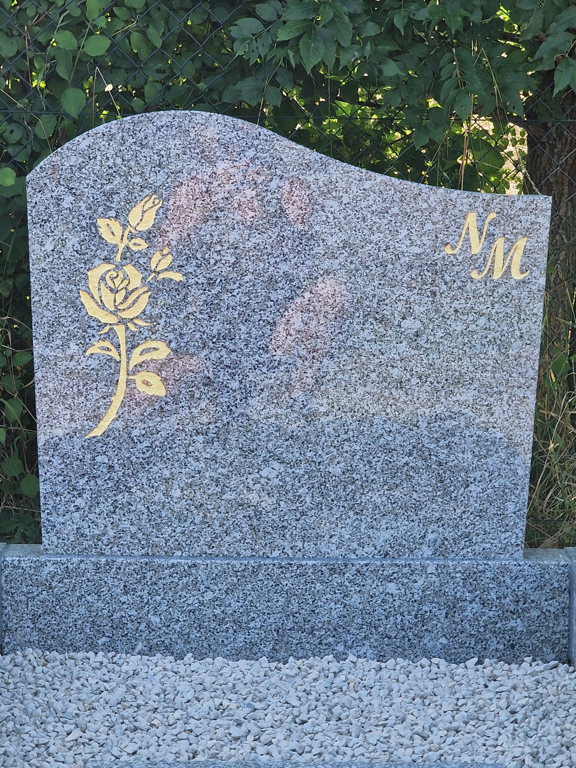 Stèle funéraire en granit gris ornée d'une rose dorée et des initiales NM, devant une haie et du gravier blanc.