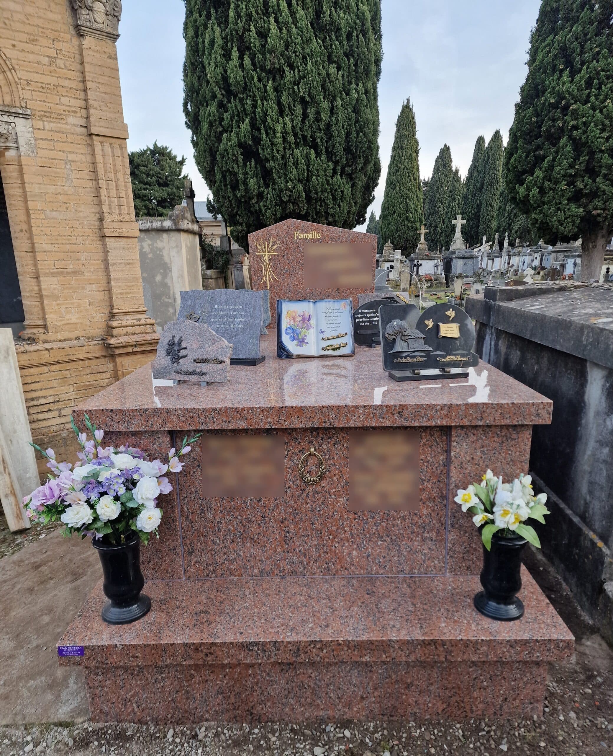 Tombe 'Famille' en granit rouge ornée de plaques commémoratives et vases de fleurs dans un cimetière arboré.