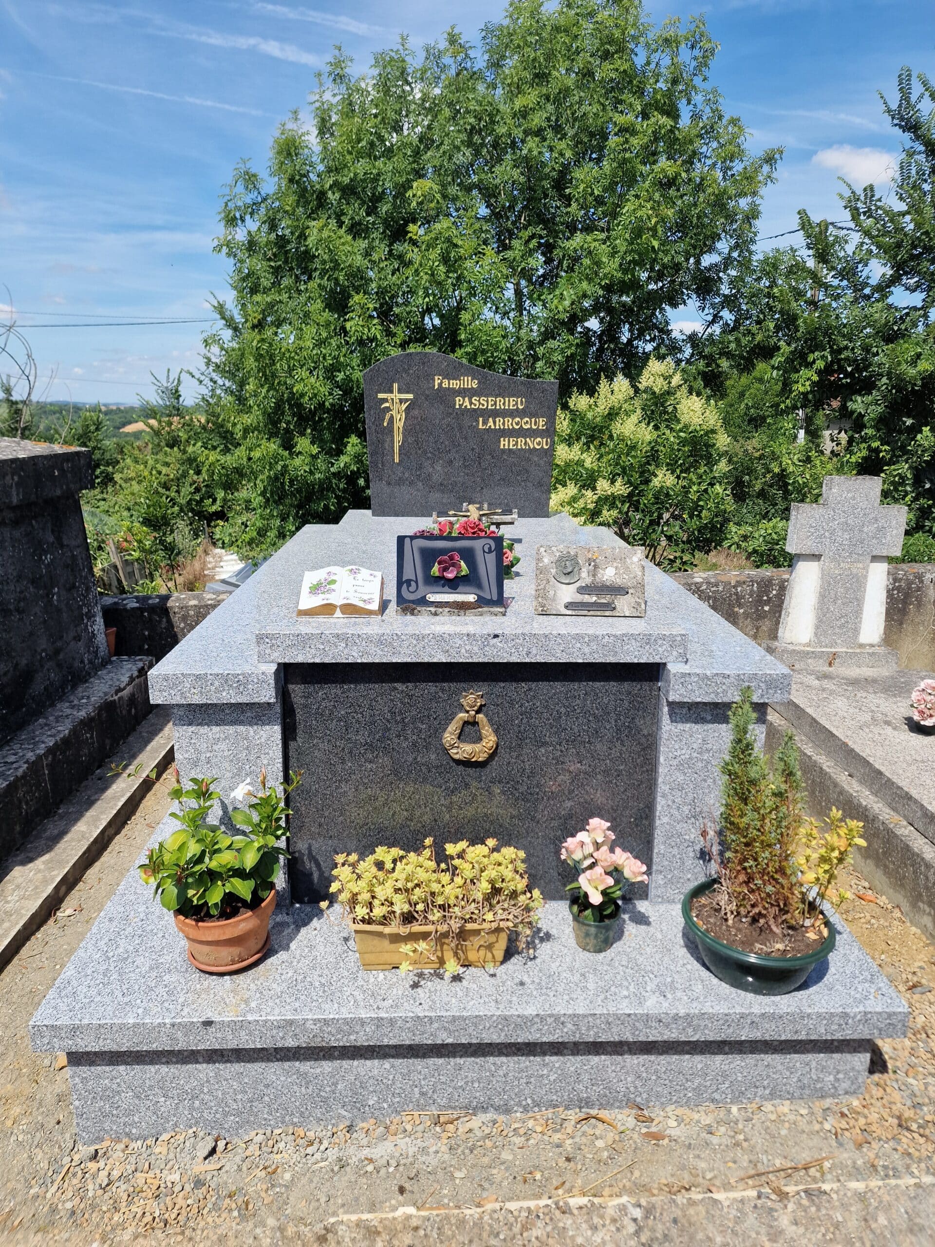 Monument funéraire Famille Passerieu Larroque Hernou, granite gris, plaques et fleurs en pots sous un ciel bleu.