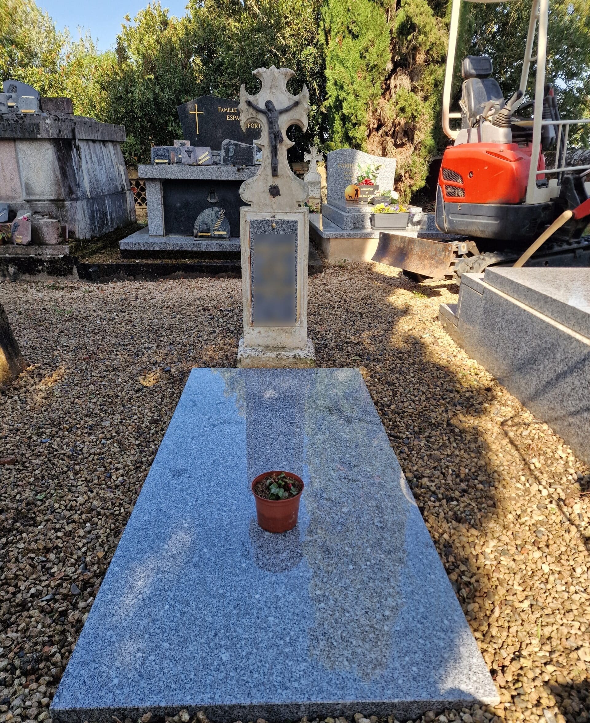 Tombe en granit gris avec croix sculptée et petite plante en pot dans un cimetière ensoleillé.