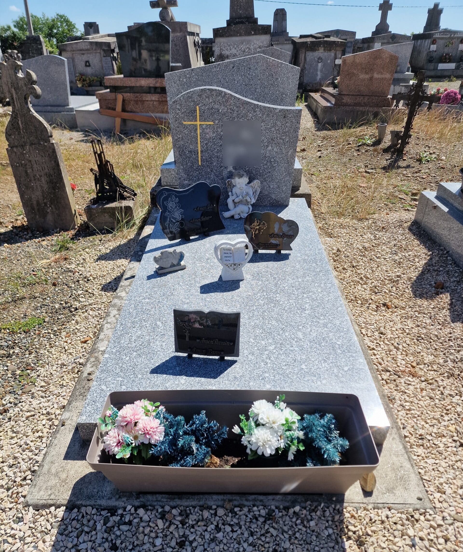 Tombe en granit gris avec croix dorée, plaques commémoratives et jardinière de fleurs dans un cimetière ensoleillé.