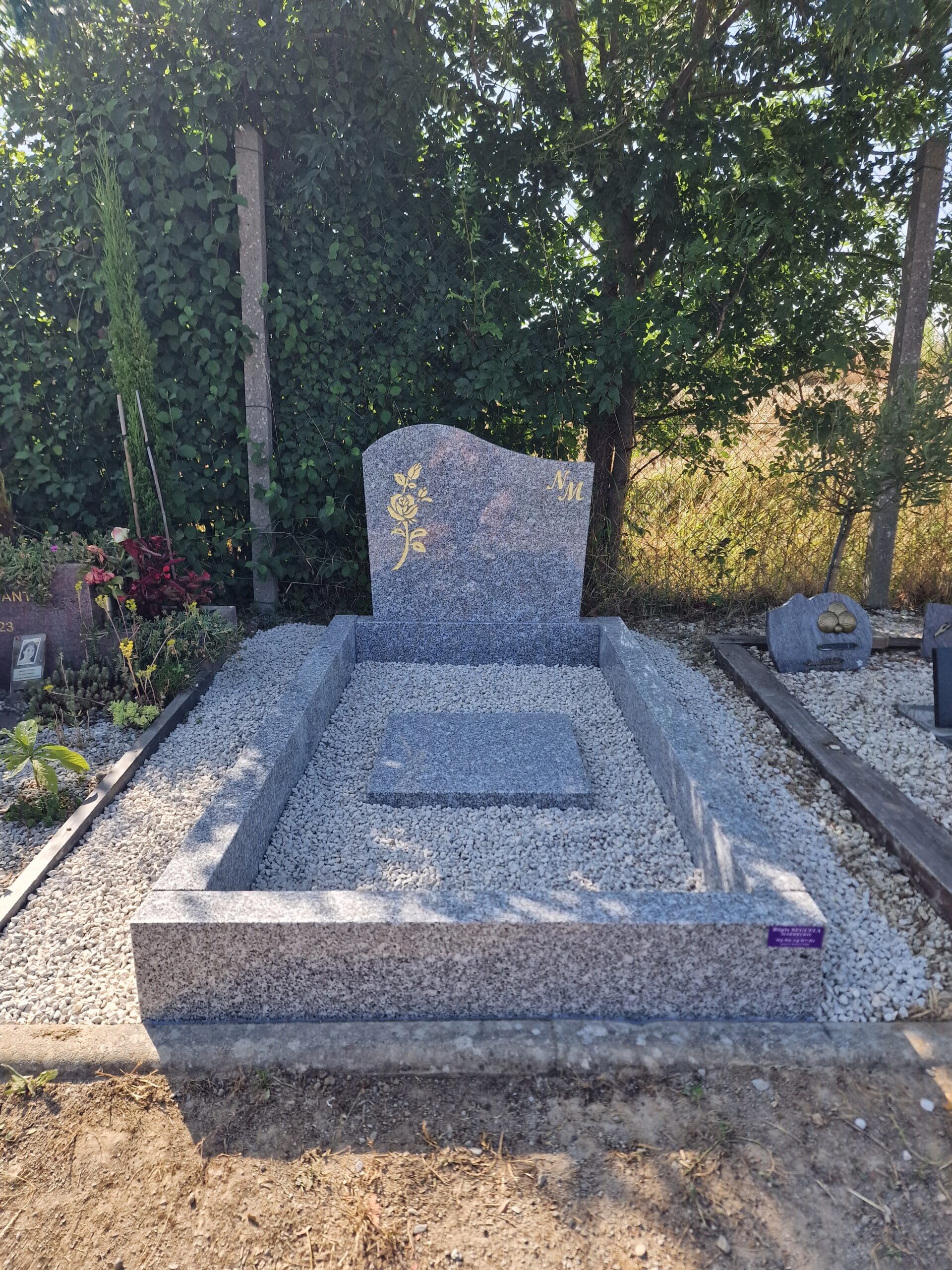 Tombe en granit gris avec rose dorée et initiales NM, remplie de graviers blancs dans un cimetière verdoyant.