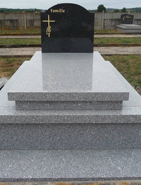 Monument funéraire : Tombe en granit noir et gris poli Tombe en granit gris et noir poli avec inscription dorée "Famille" et croix feuillue. Cimetière avec murs en béton en arrière-plan.