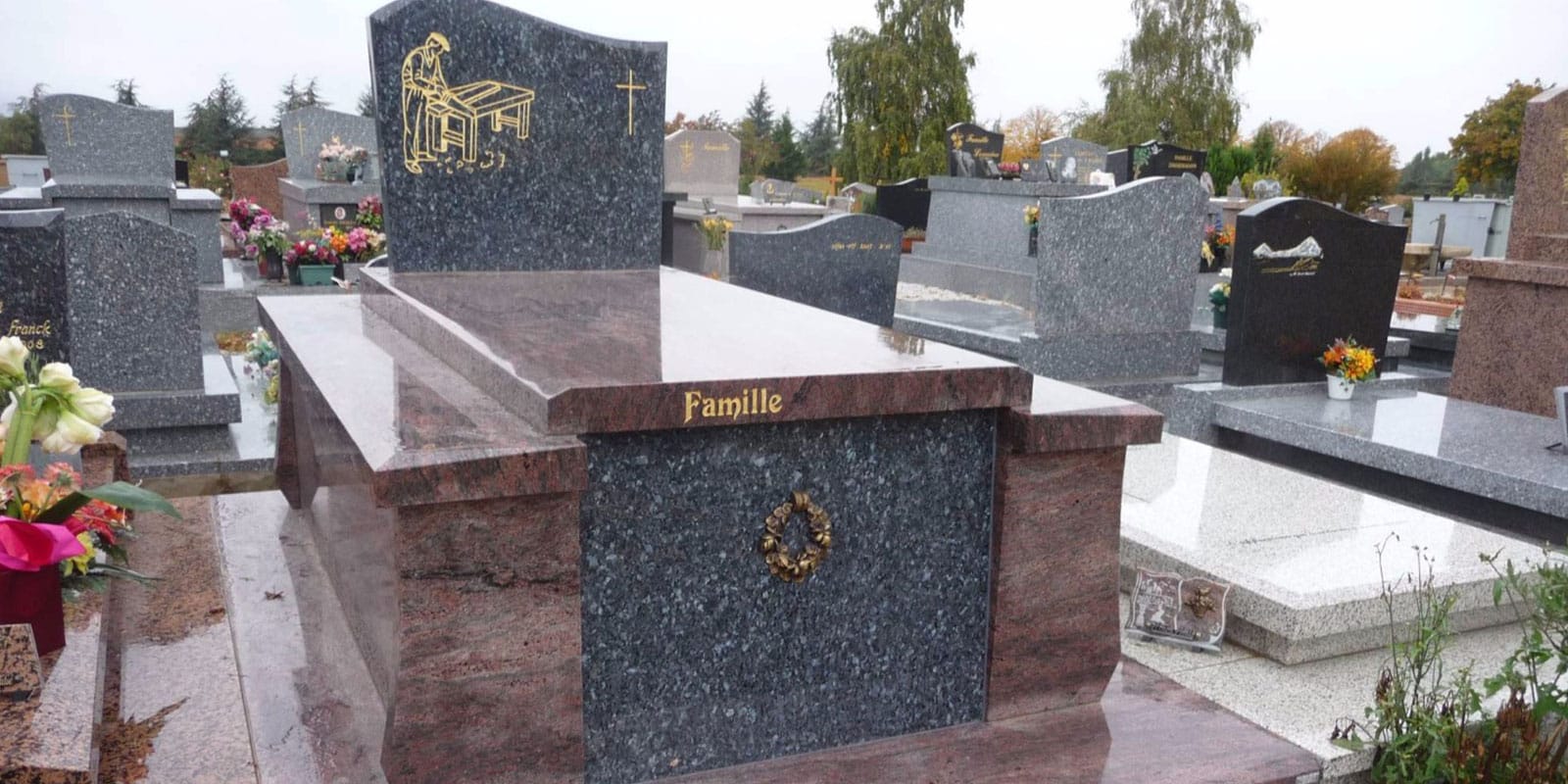 Grande tombe en granit gris et rouge au cimetière. Gravure dorée d'un artisan, croix, texte "Famille" et couronne. Plusieurs autres monuments sont visibles.