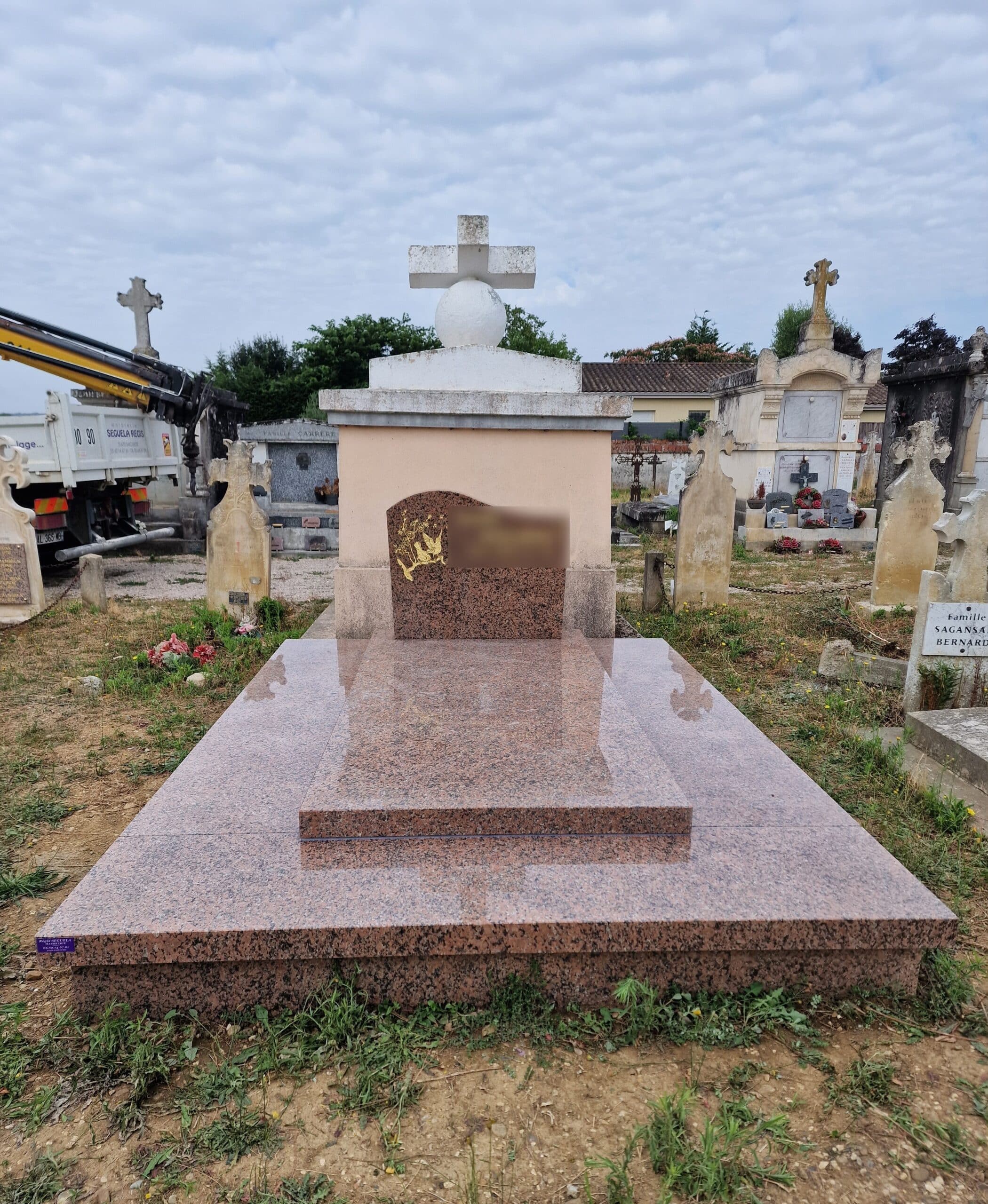 Tombe en granite rose poli avec gravure dorée devant un monument à croix blanche dans un cimetière.