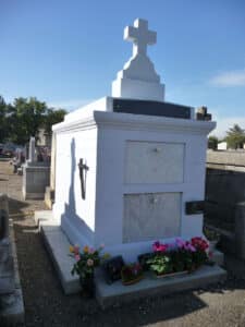 Caveau funéraire blanc, de style classique, coiffé d'une grande croix, décoré de fleurs, dans un cimetière sous un ciel bleu.