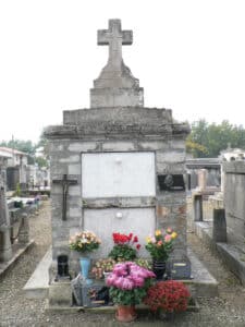 Caveau funéraire en pierre grise avec une croix au sommet, orné d'une crucifixion et de bouquets de fleurs colorées devant les plaques tombales où l'on distingue 'DEGRETS'.