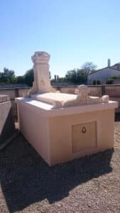 Monument funéraire en pierre claire, style mausolée avec colonne crénelée et ornementations sculptées, devant un mur et des arbres sous un ciel bleu.