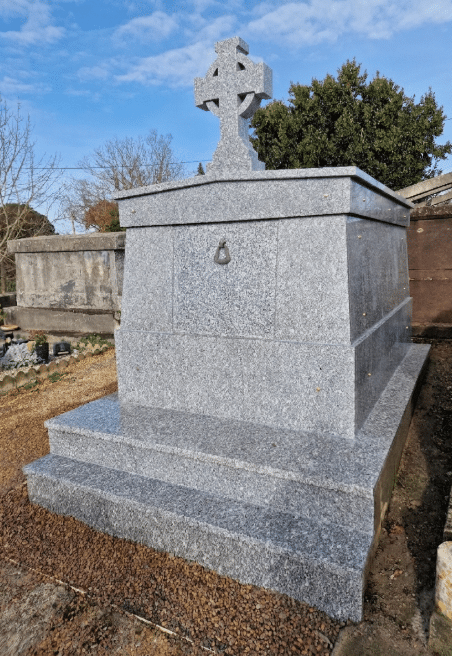 Monument funéraire en granit gris surmonté d'une croix celtique sculptée dans un cimetière sous un ciel bleu.