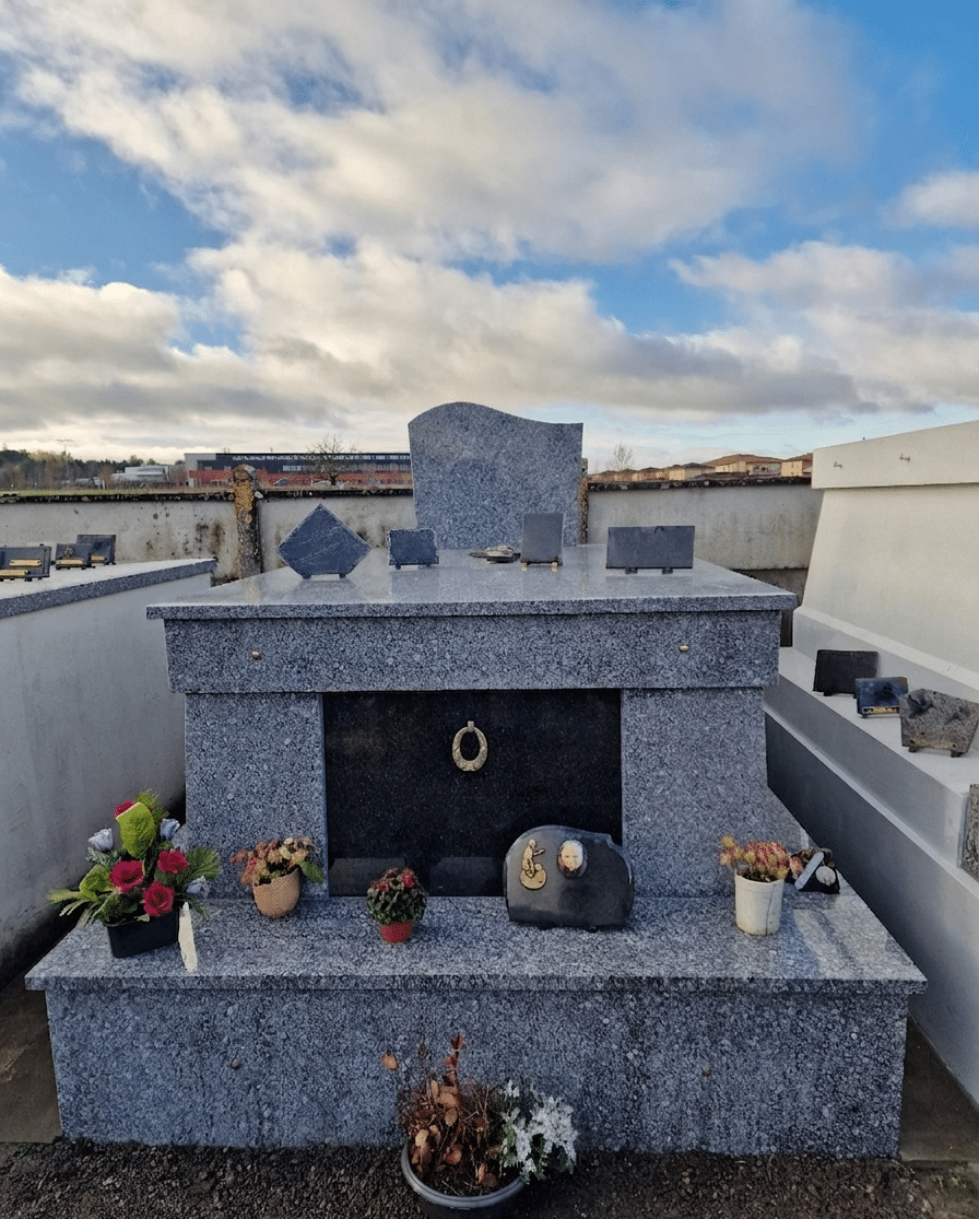 Monument funéraire en granit bleu orné de plaques souvenirs et de fleurs sous un ciel nuageux de cimetière.