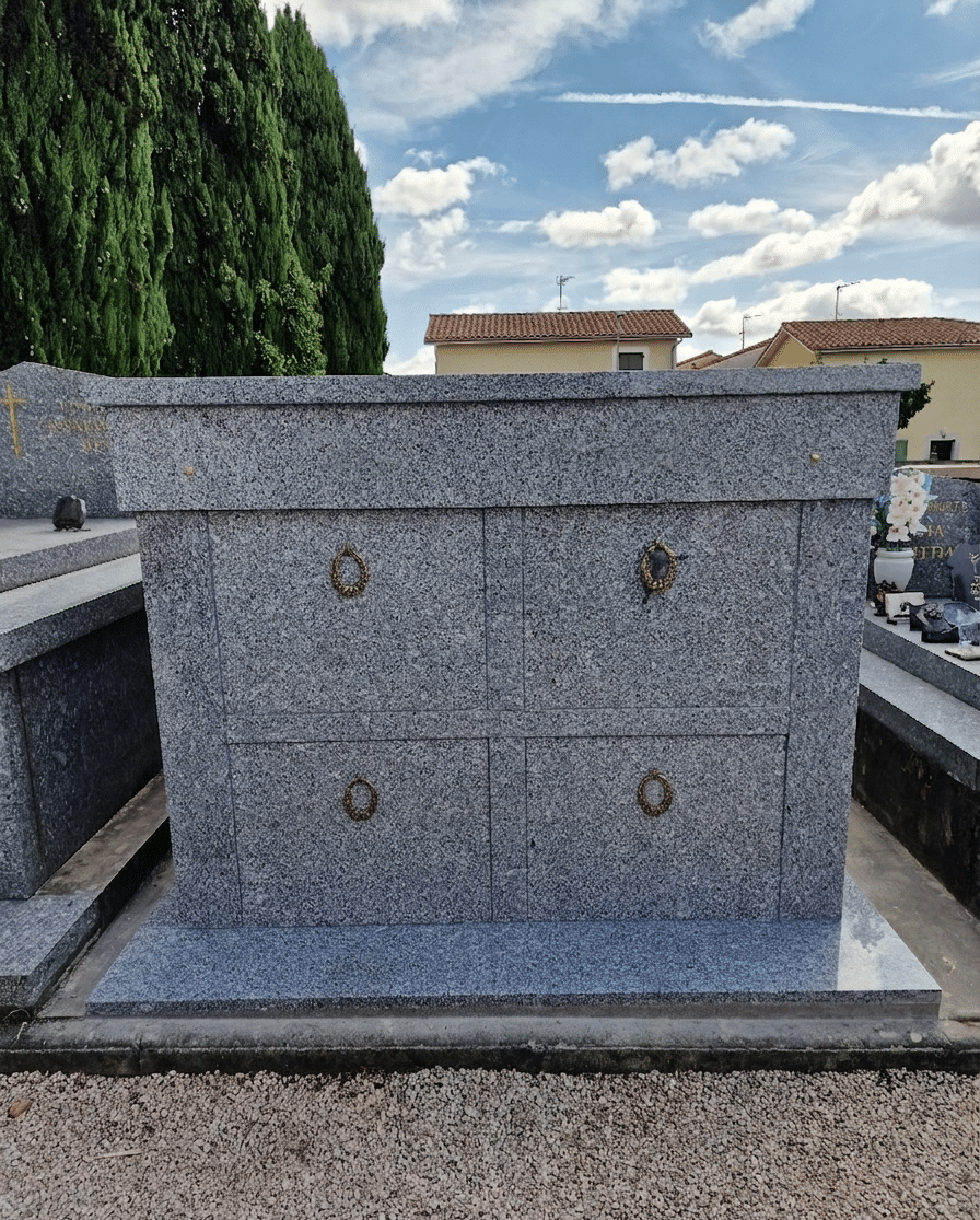 Monument funéraire en granite gris avec quatre niches et anneaux dorés dans un cimetière.