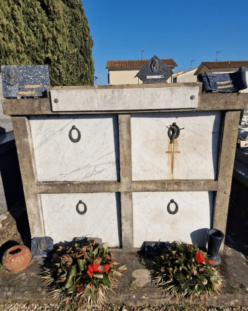 Mausolée en marbre blanc à quatre niches avec crucifix, plaques funéraires et couronnes de fleurs sous un ciel bleu.