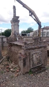 Sépulture de famille délabrée, partiellement en briques, avec une stèle ornementée portant l'inscription 'FAMILLE' et un crucifix, en cours de manutention par une grue.